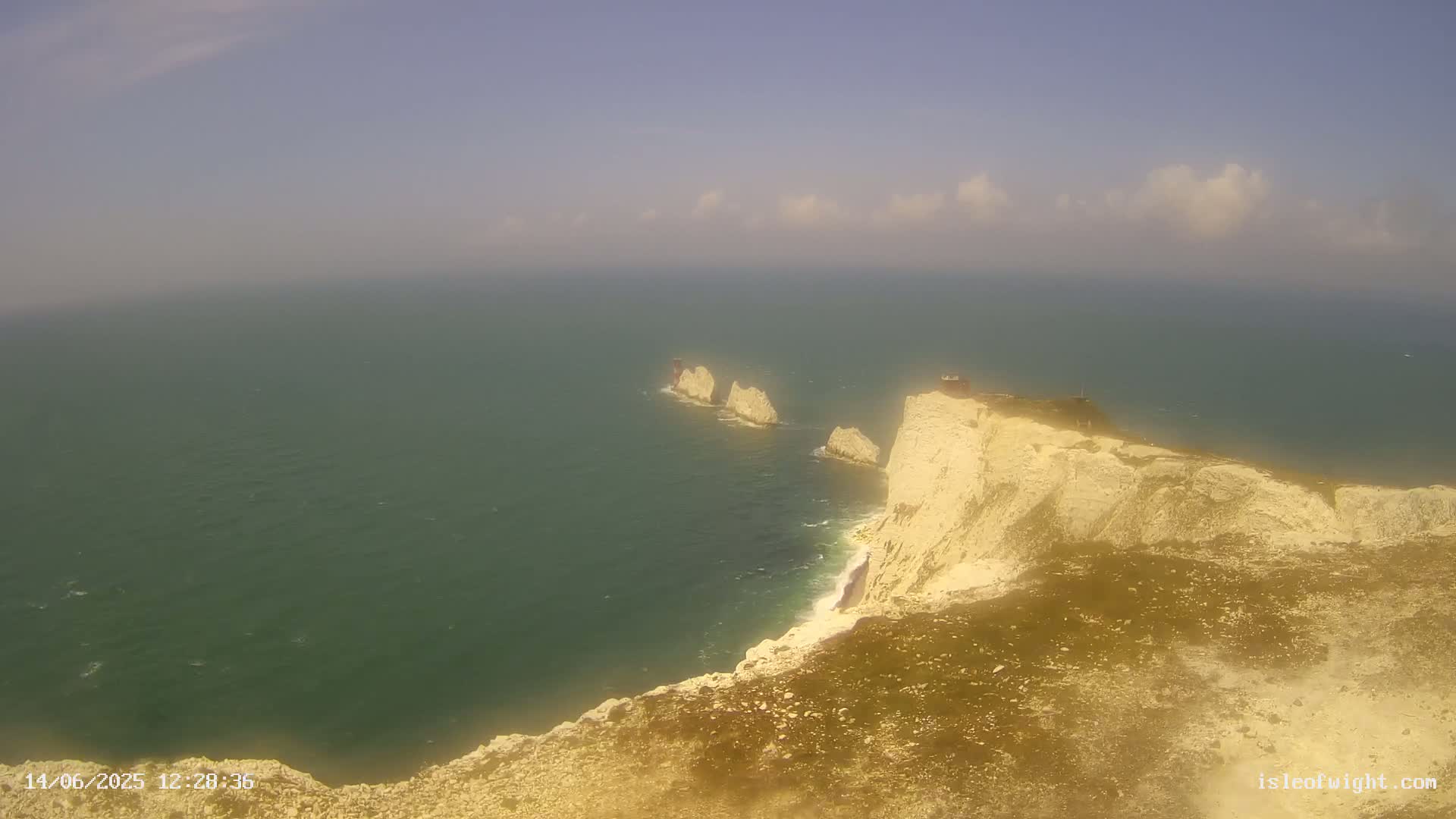 A high-angle view shows white cliffs meeting a calm, greenish-blue sea with several smaller rock formations jutting from the water.