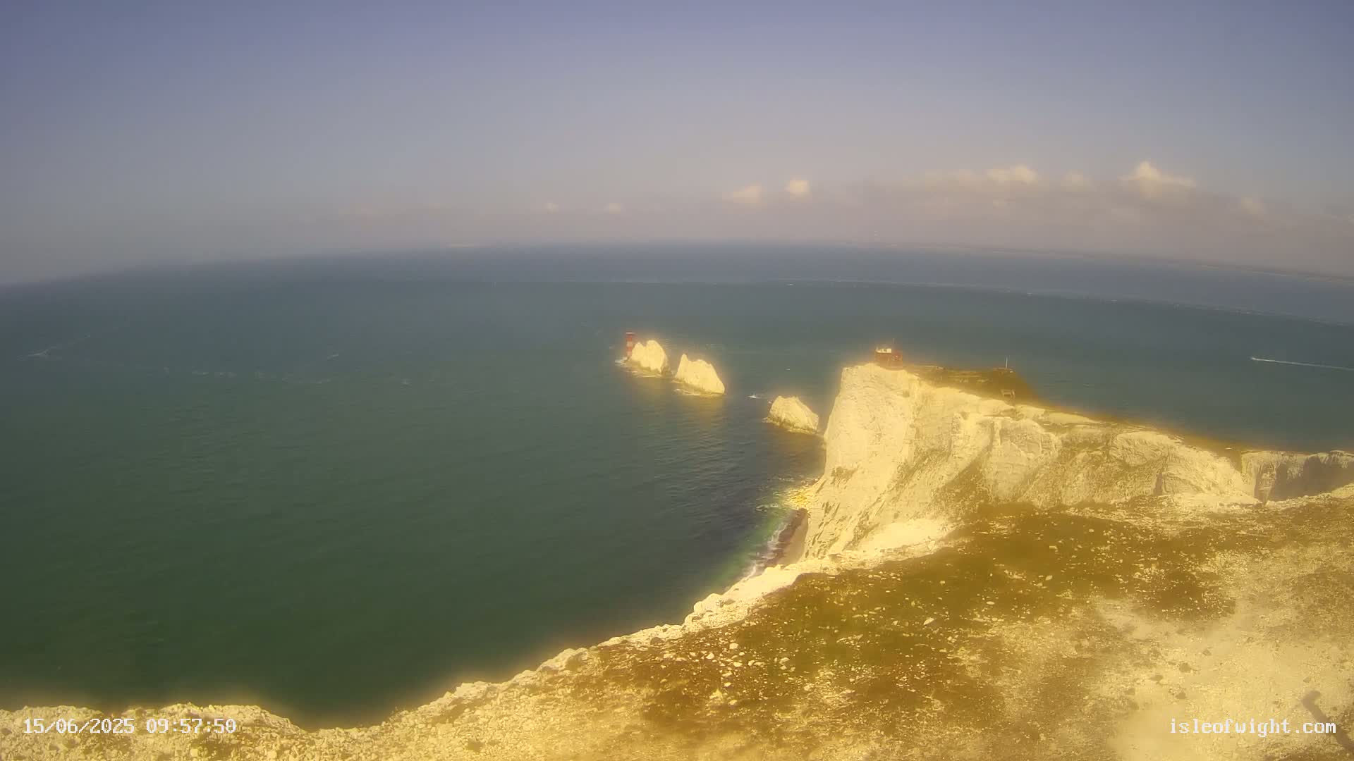 A high-angle view shows a bright, sunny day overlooking a dramatic white cliffside overlooking a calm, dark-green sea with several small rocky islands.