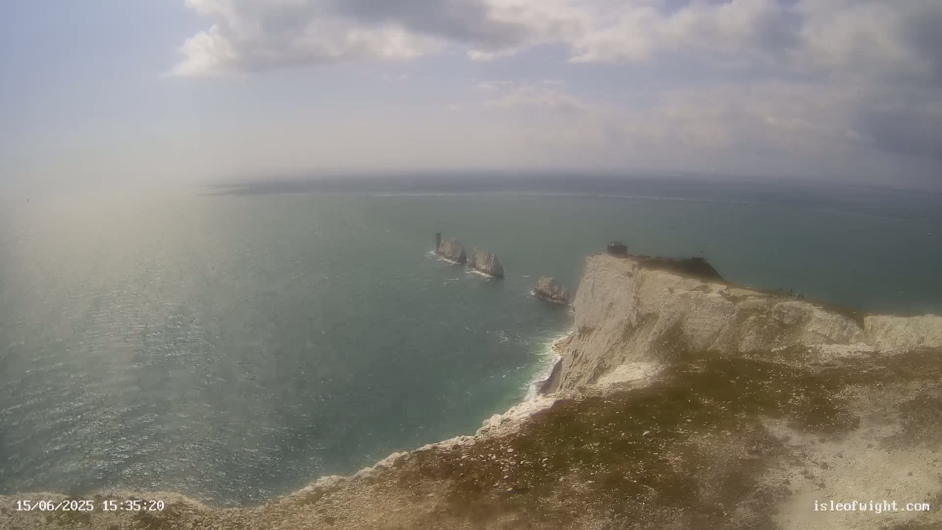 A high-angle view shows a rocky, white cliff overlooking a calm, hazy sea with several small islands in the distance under a partly cloudy sky.