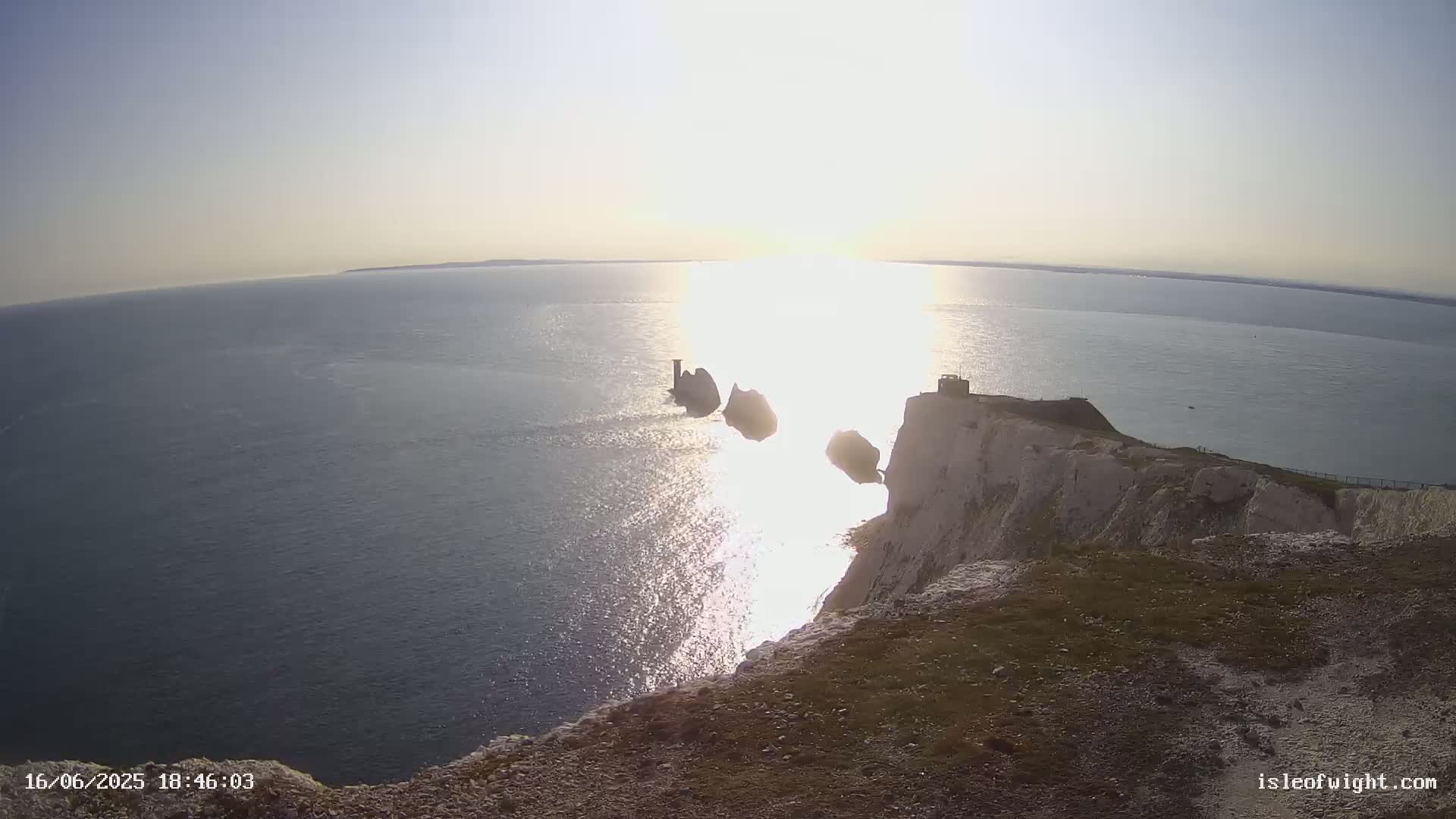 A bright, sunny day shows a view from a clifftop overlooking the ocean, featuring several rock formations and a small structure on the cliff edge.