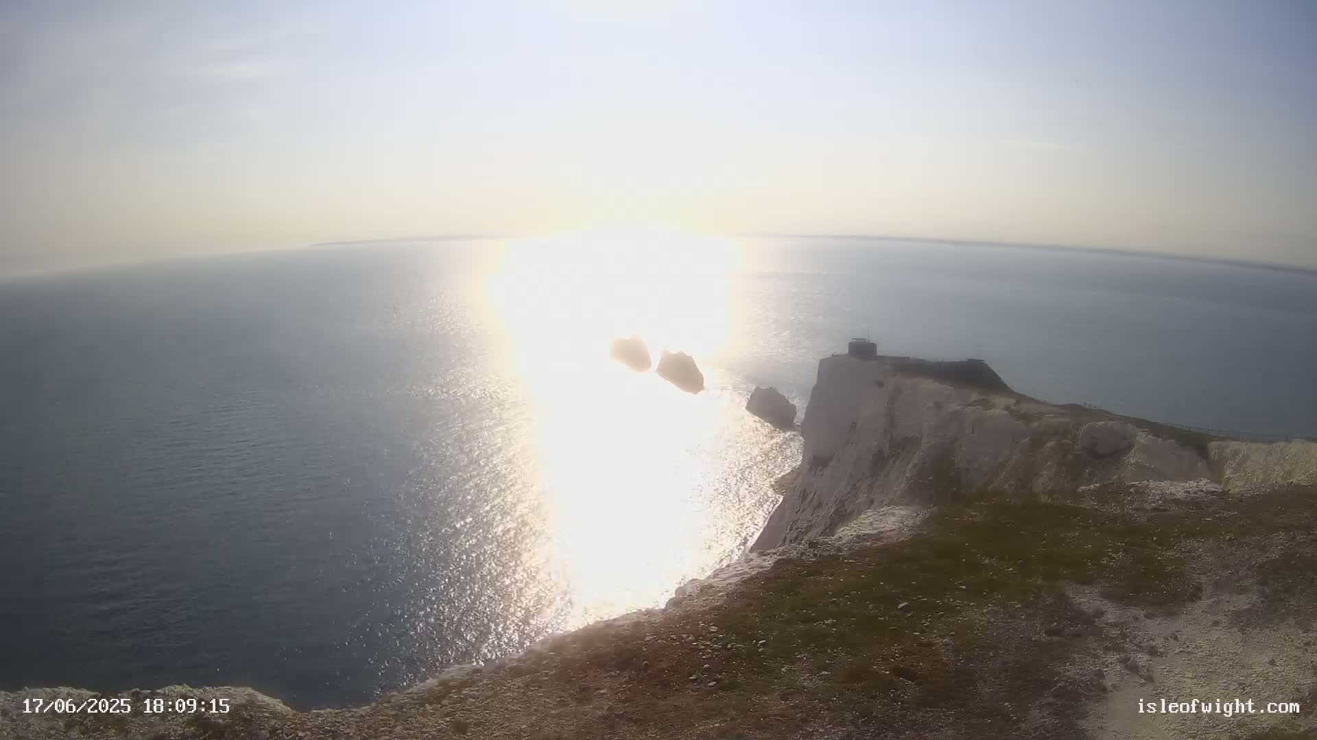 A bright, sunny day shows a view from a cliff overlooking the ocean, with two small islands and a building visible in the distance.