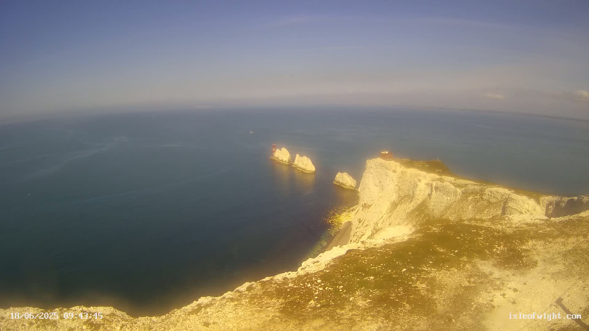 A high-angle view shows white cliffs meeting a calm, blue ocean, with several smaller white rock formations just offshore under a clear, bright sky.