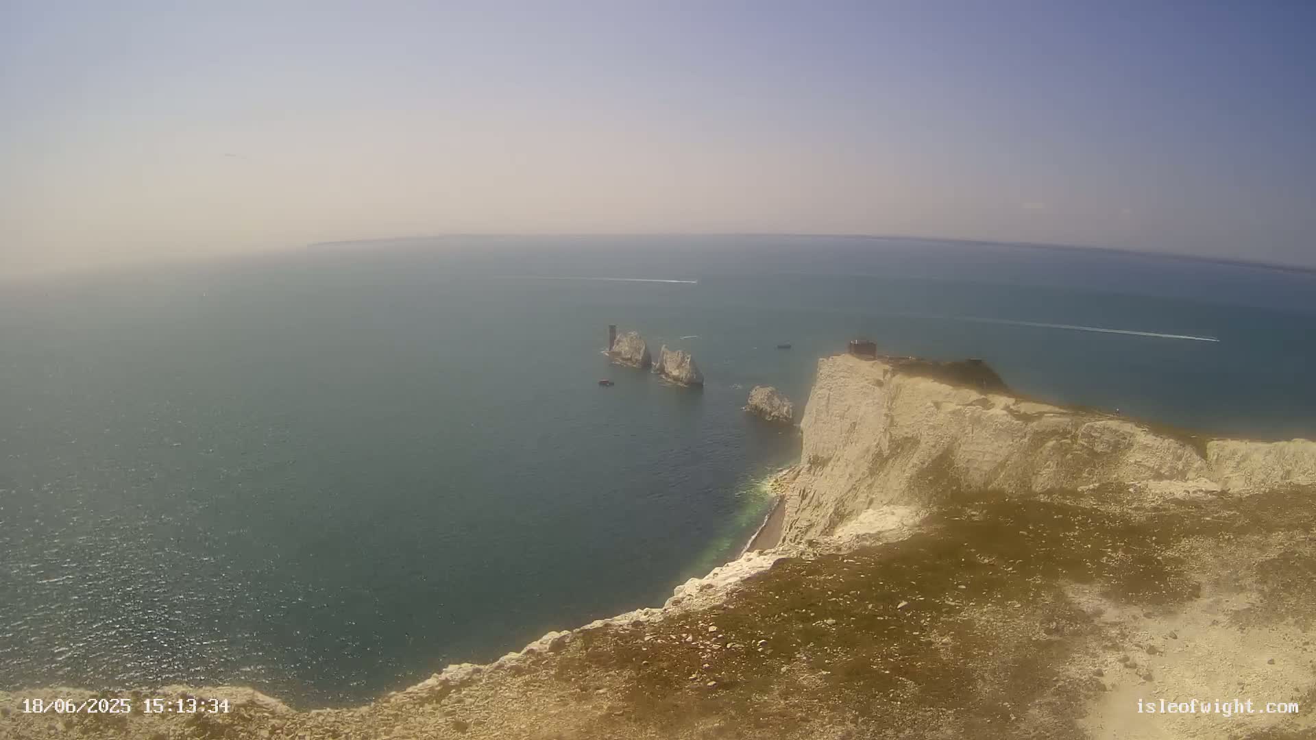 A hazy, sunny day reveals a high-angle view of a white cliff overlooking the ocean, where several small rocky islands and a few boats are visible in the distance.
