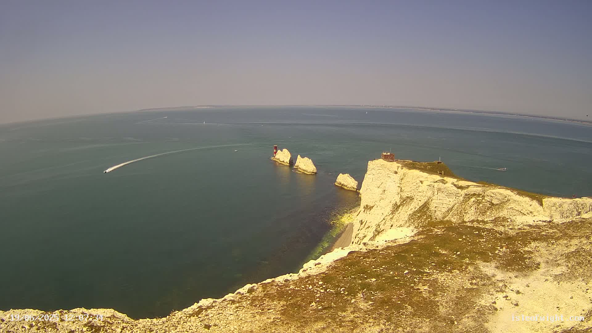 A high-angle view shows white cliffs overlooking a calm, dark-blue sea where several stacks of rock jut out from the water on a sunny day.