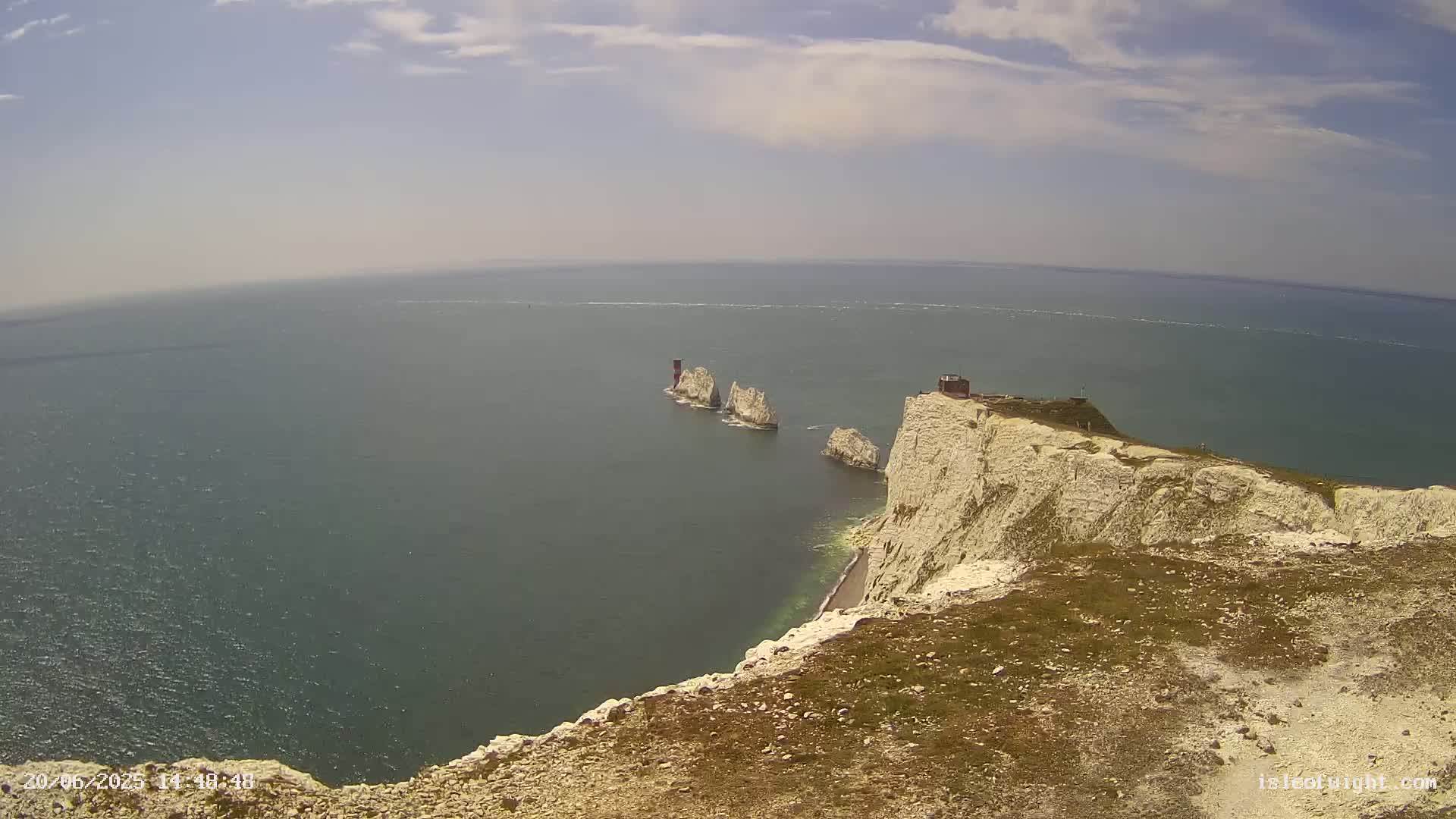 A high-angle view shows a rocky, white cliff overlooking a calm sea with three small, distant islands on a partly sunny day.