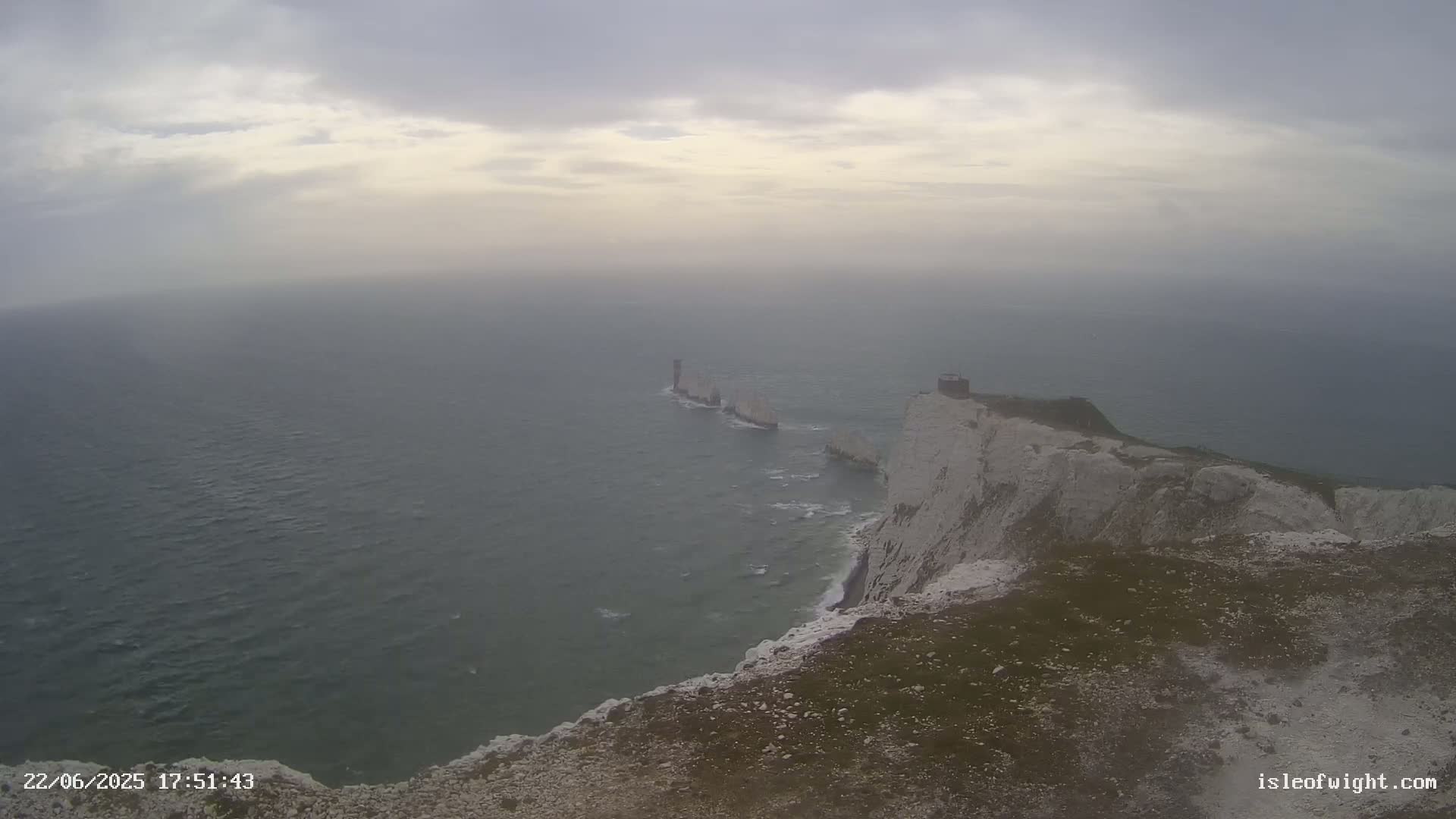 A hazy view of white cliffs and a chain of rocks extending into a grey sea under a cloudy sky.