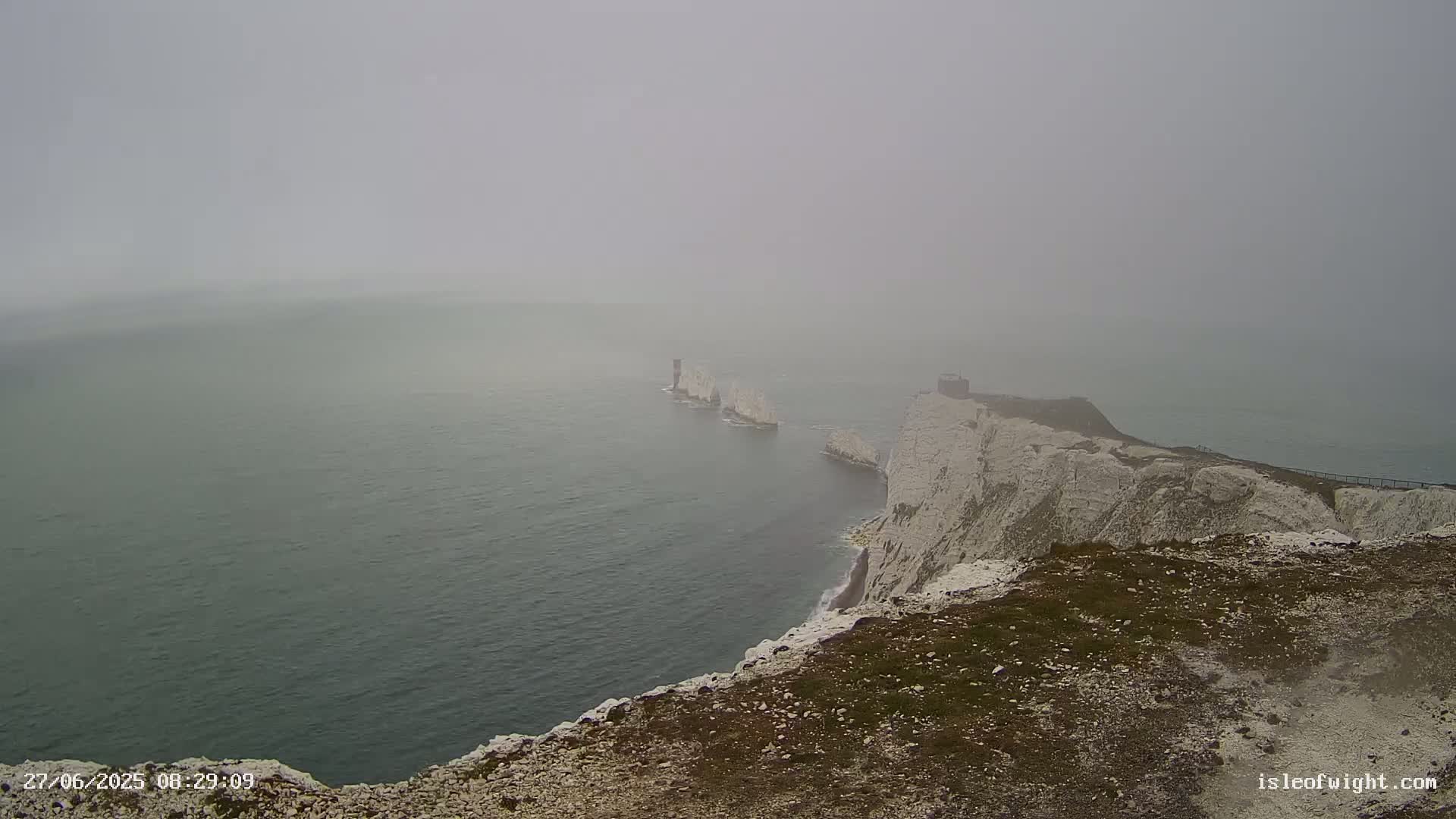 A foggy view from a clifftop overlooking a body of water with several distant white rock formations.