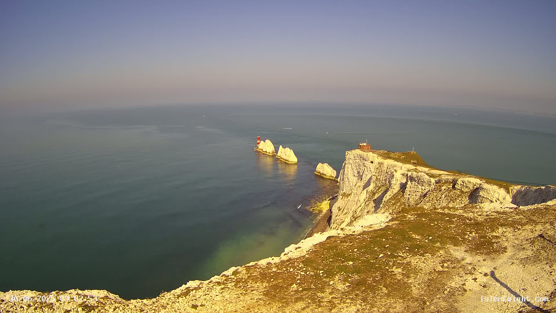 White cliffs overlook a calm sea with several white rock formations and a small red structure on one of them.