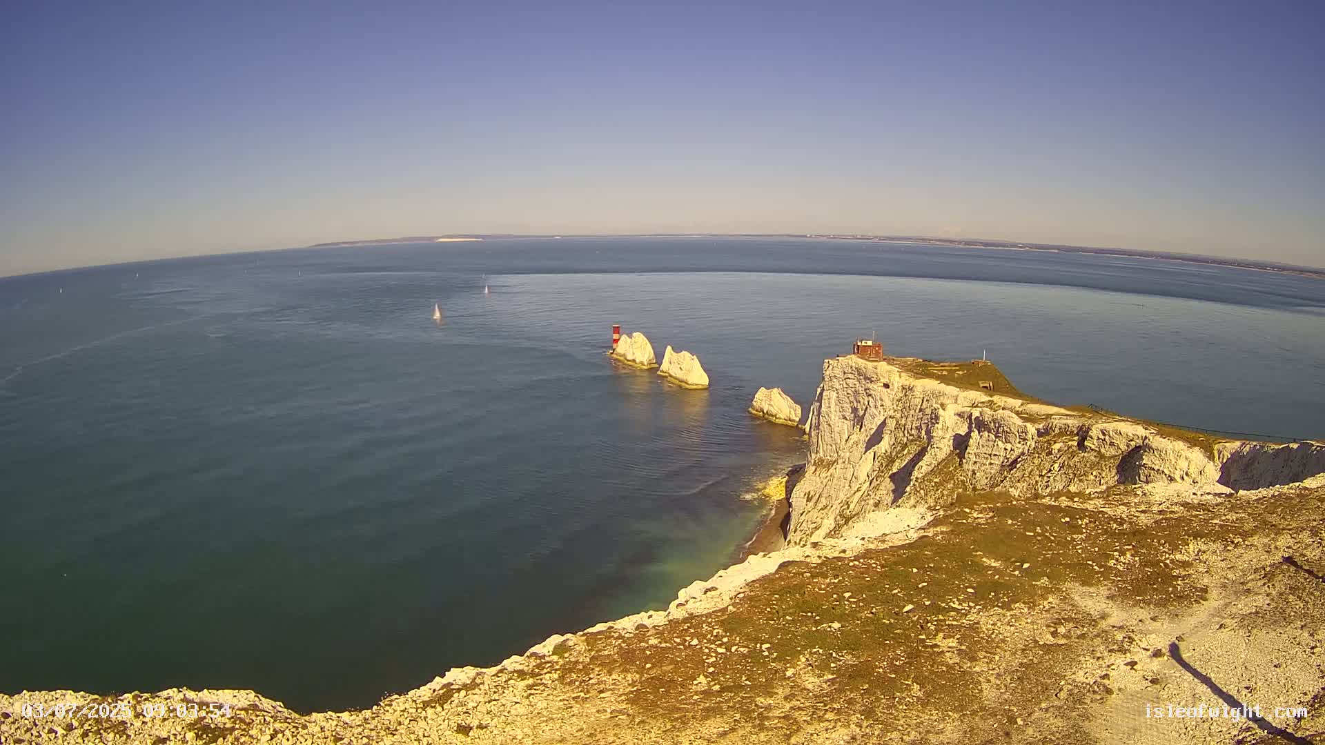 A high-angle view shows a white cliff overlooking a body of water with several small islands and sailboats under a clear blue sky.