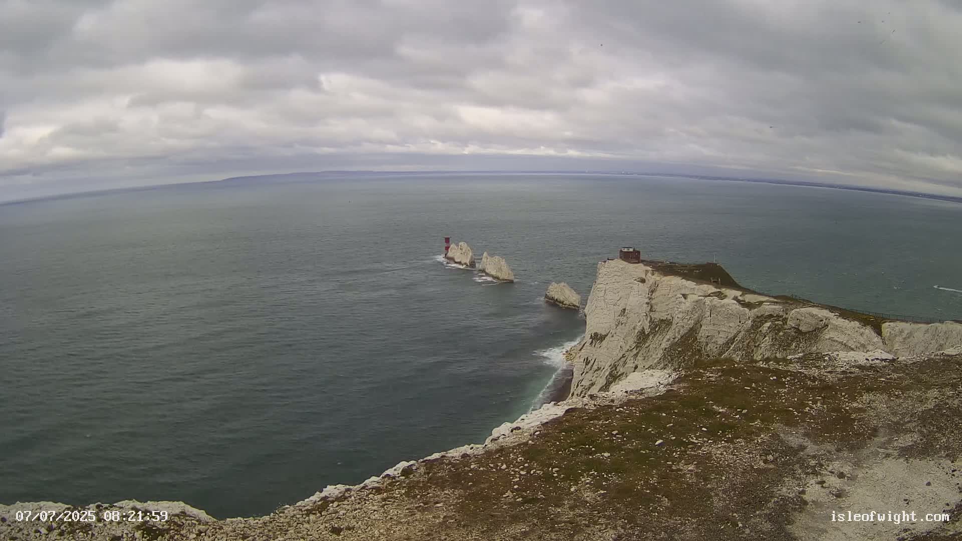 White cliffs overlooking a dark-grey sea with three small white rock formations and a small structure in the distance under an overcast sky.