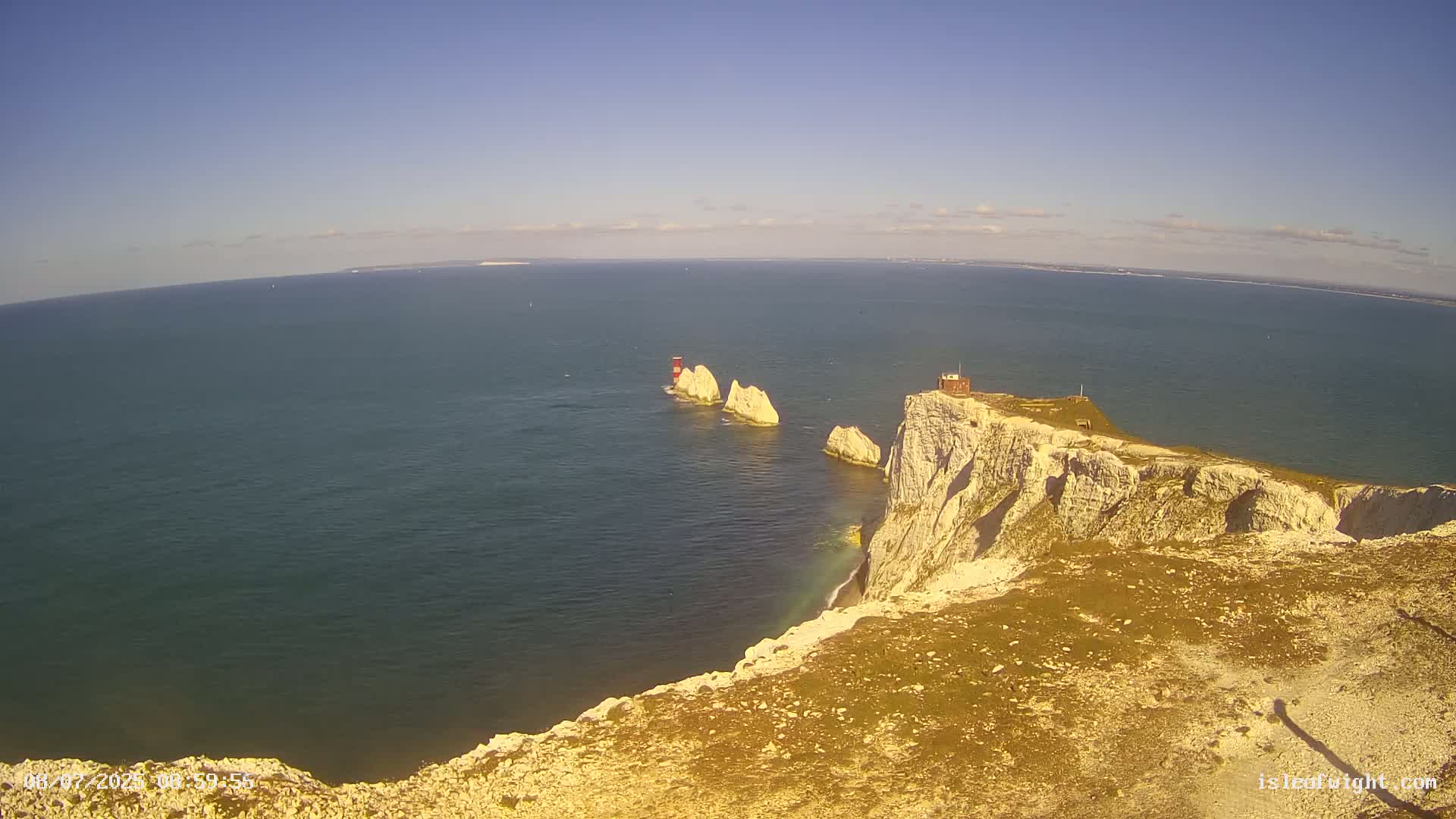 A high-angle view shows a rocky, white cliff overlooking a calm, blue ocean; in the distance are several smaller white rock formations and a lighthouse.