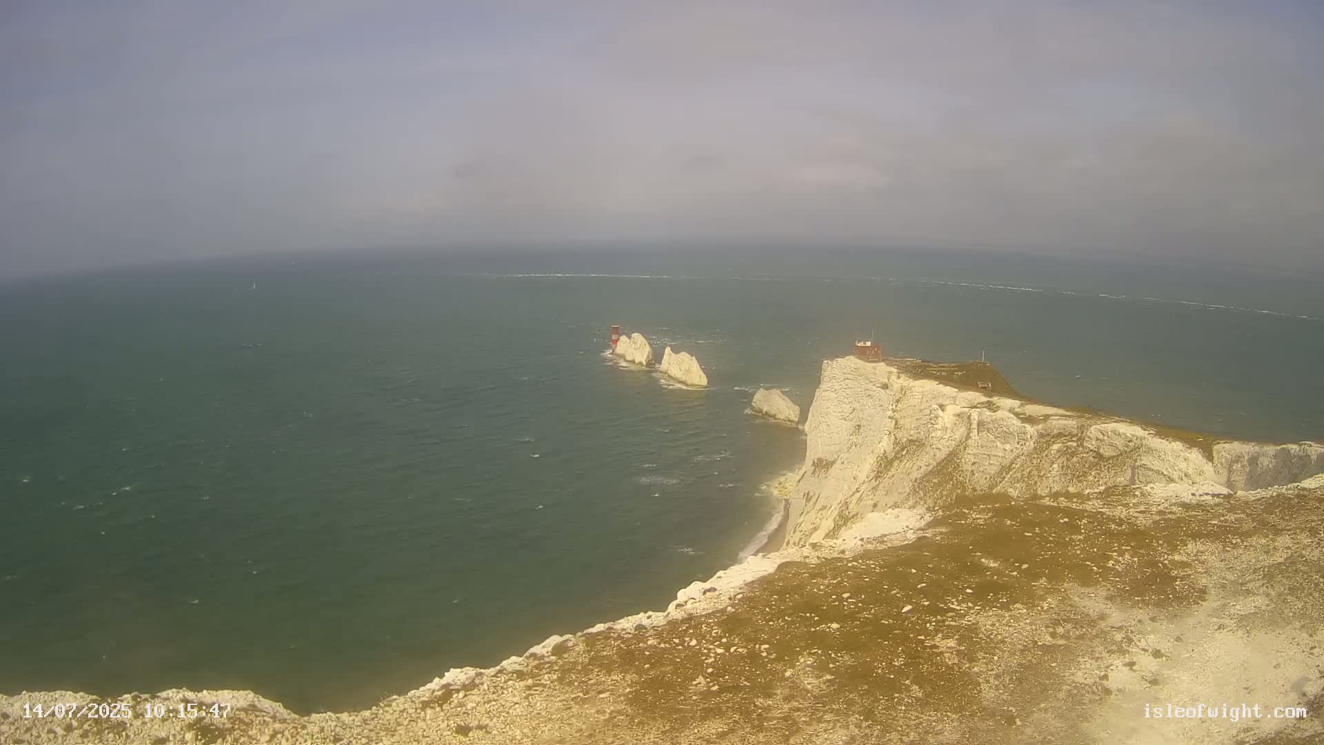 A high-angle view shows a rocky, white cliff overlooking a green sea with a few small, white rock formations and a small red building visible in the distance under a slightly hazy sky.