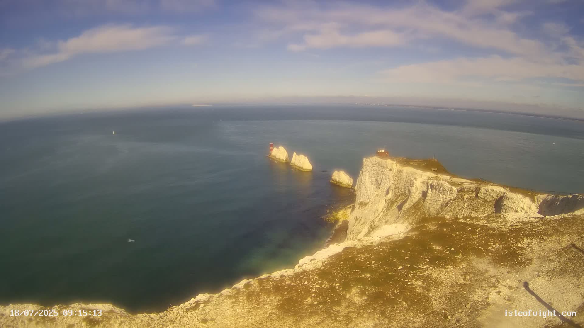 White cliffs overlook a calm, teal ocean with three smaller white rock formations and a small red structure visible in the distance under a mostly clear, sunny sky.