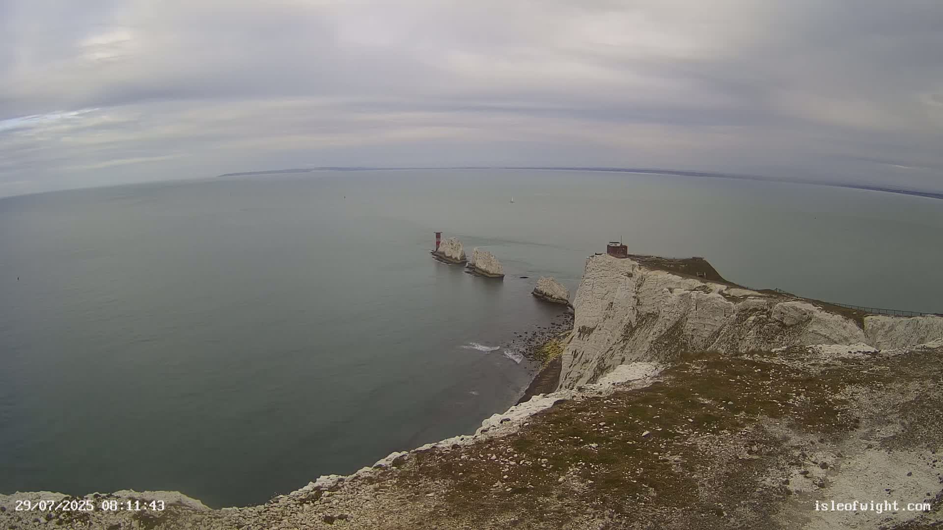 A high-angle view shows a rocky coastline with white cliffs extending into a calm, gray sea, featuring several small, distant islands with a small structure on one of them, under an overcast sky.