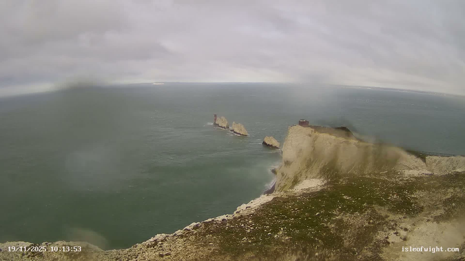 A dramatic coastal scene depicts white chalk cliffs and the iconic sea stacks known as The Needles amidst choppy green water under a heavily overcast and possibly rainy sky.