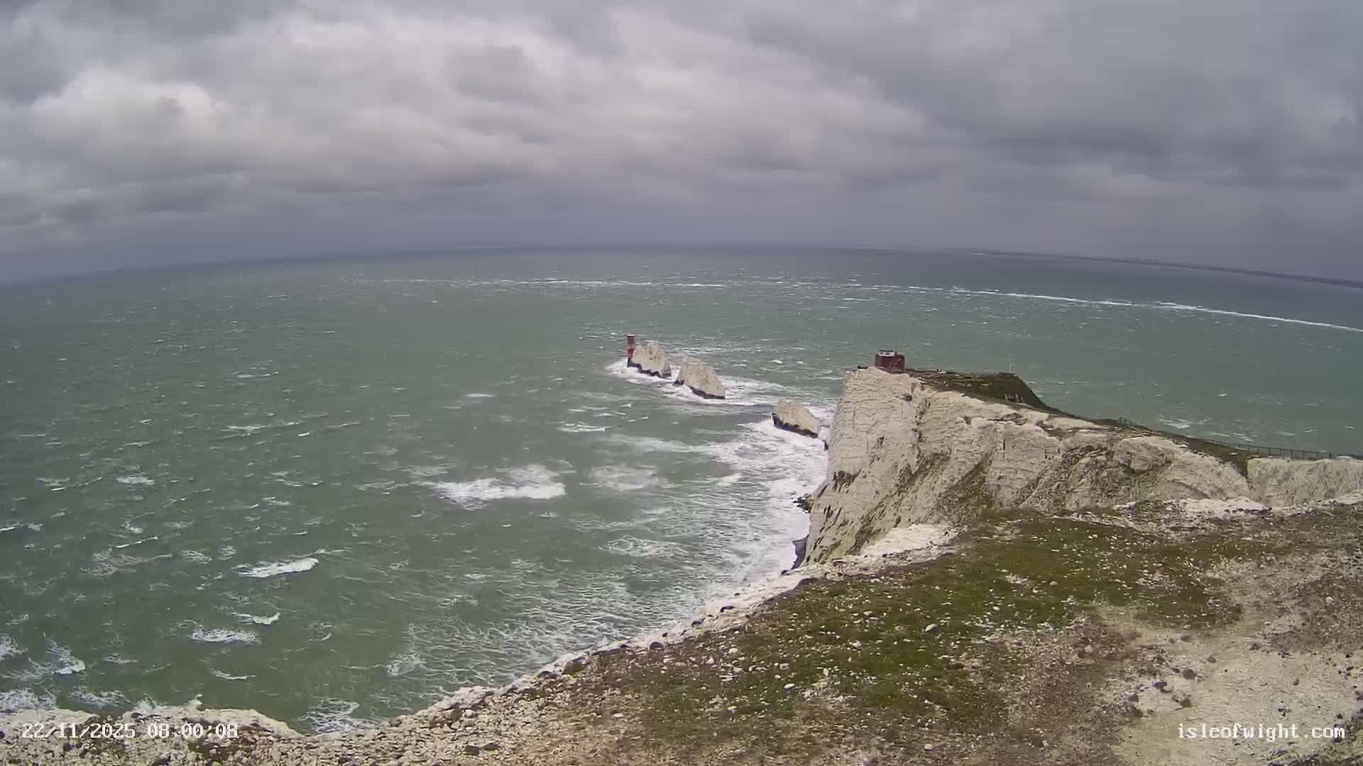 A dramatic coastal scene shows white chalk cliffs and the iconic Needles rock formations with their lighthouse emerging from a choppy green-grey sea under a heavy, overcast sky.