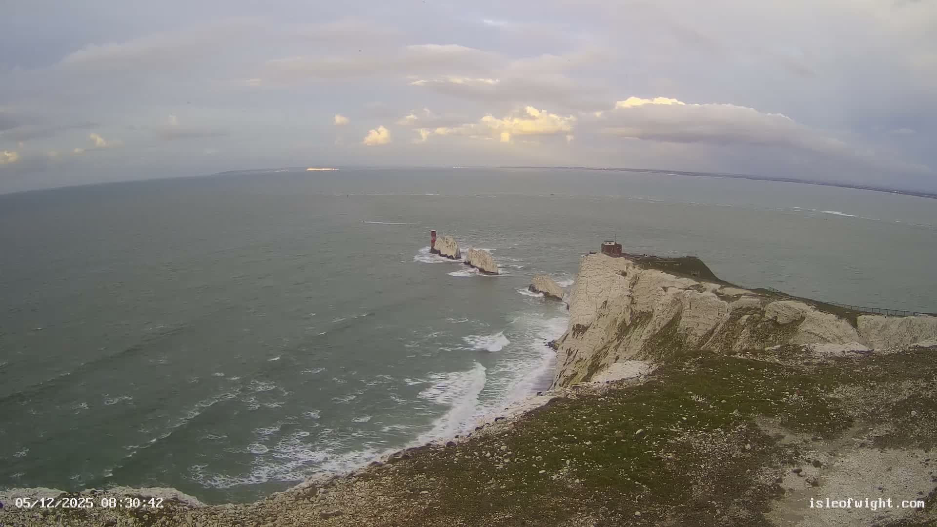 An expansive view captures the iconic Needles sea stacks and lighthouse off the Isle of Wight's white cliffs, with rough waves breaking against the shore under a mostly cloudy sky.