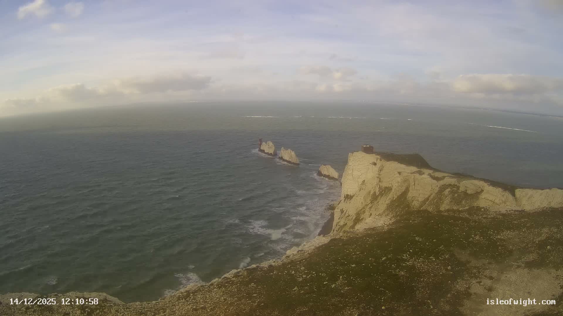 An expansive view captures the iconic Needles sea stacks and lighthouse off the Isle of Wight's white cliffs, with rough waves breaking against the shore under a mostly cloudy sky.