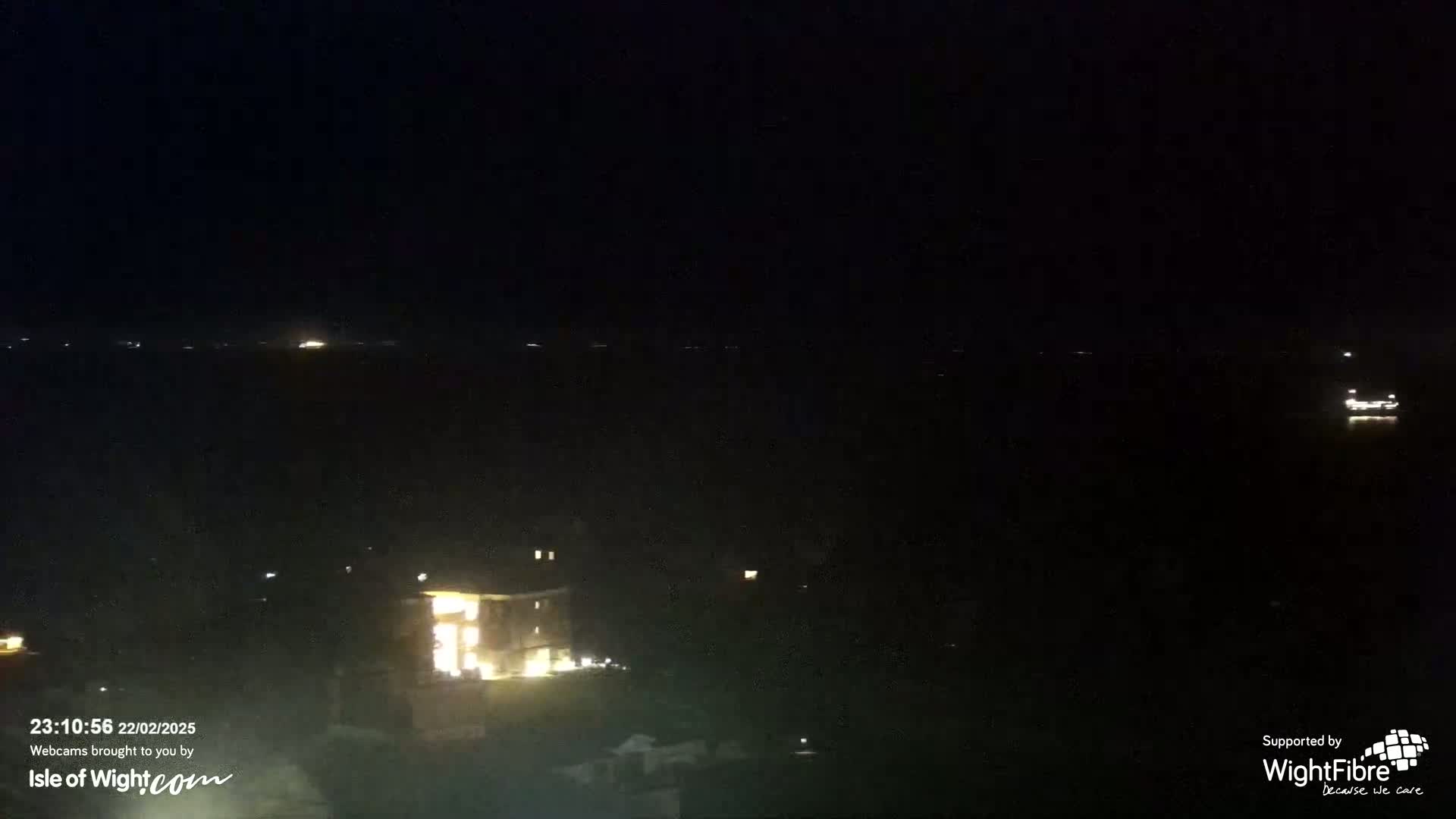 A nighttime long shot shows a dark body of water with a few distant lights, and a brightly lit building close to the bottom of the frame.