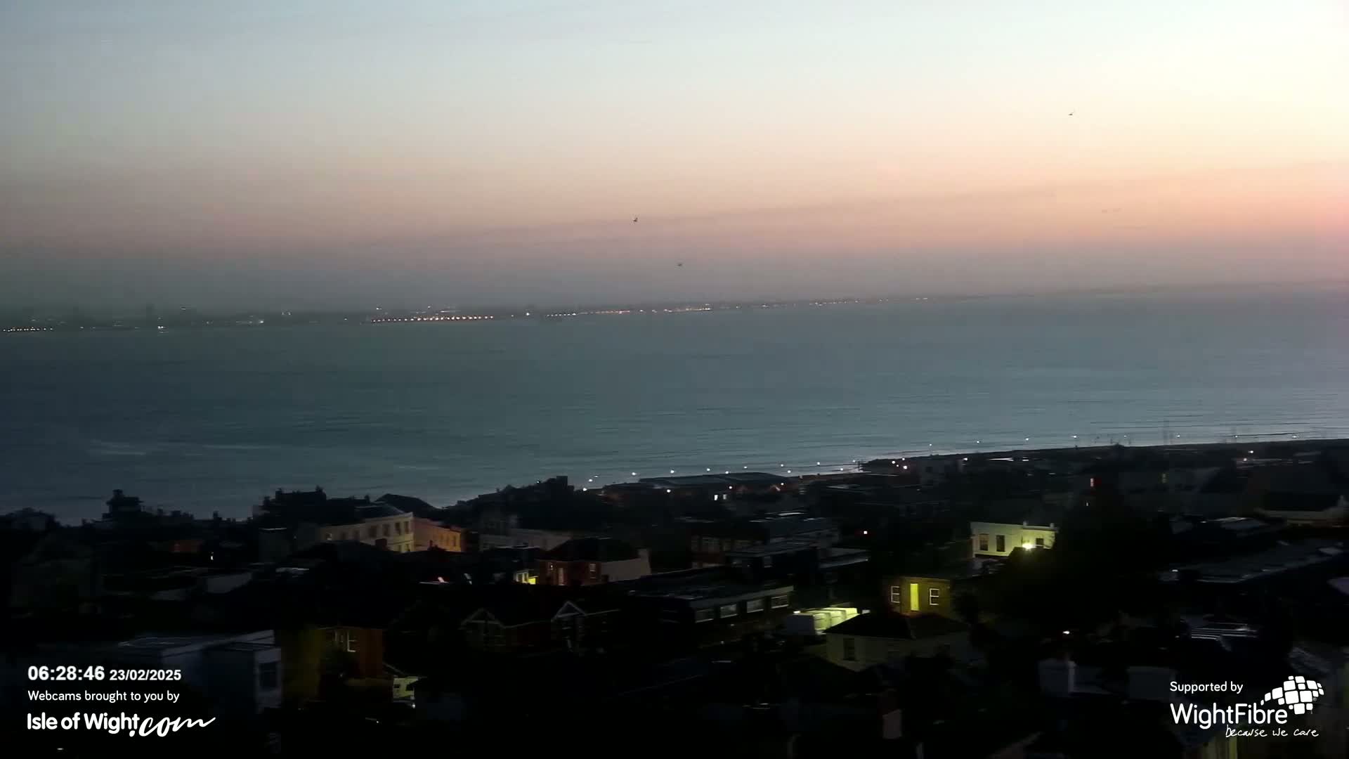A coastal town at twilight is shown, with a calm sea extending to a distant shoreline under a pinkish sky.