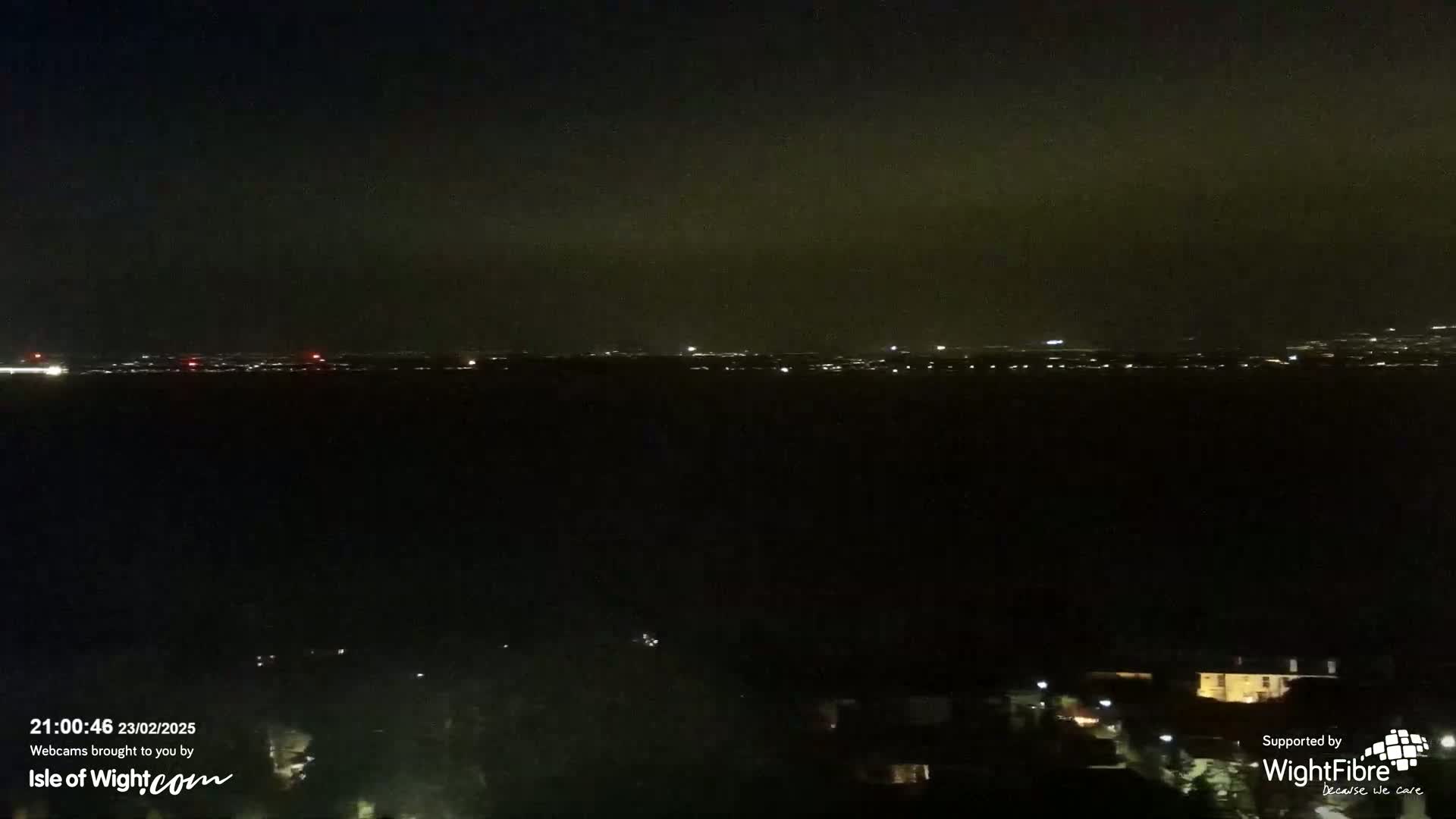 A nighttime long shot shows a distant city skyline faintly lit against a dark, cloudy sky, with a few lights visible closer to the foreground.