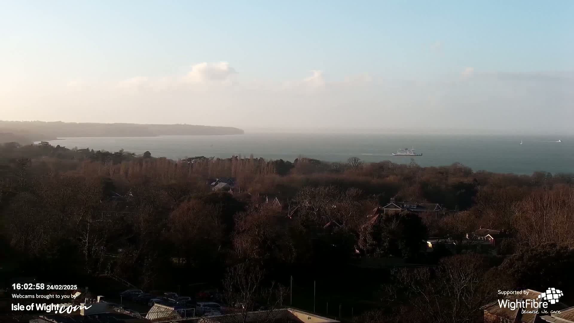 A tranquil coastal scene on a partly cloudy day shows a body of water with a ferry and sailboat in the distance, overlooking a wooded area with residential buildings.