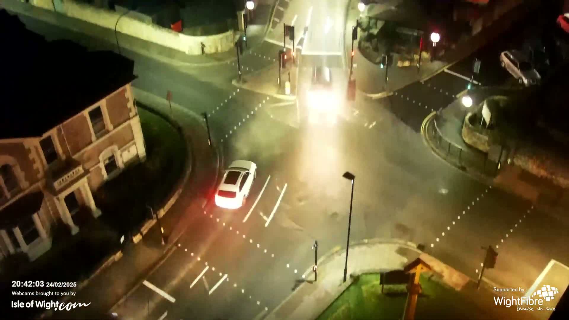 A white SUV drives through a wet, nighttime intersection near a multi-story building.