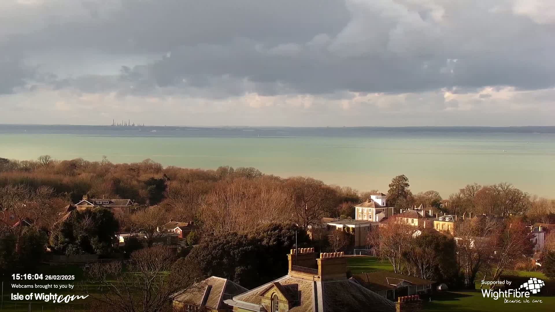 A view of a calm, light-green body of water under a mostly cloudy sky, with bare winter trees and buildings in the foreground.