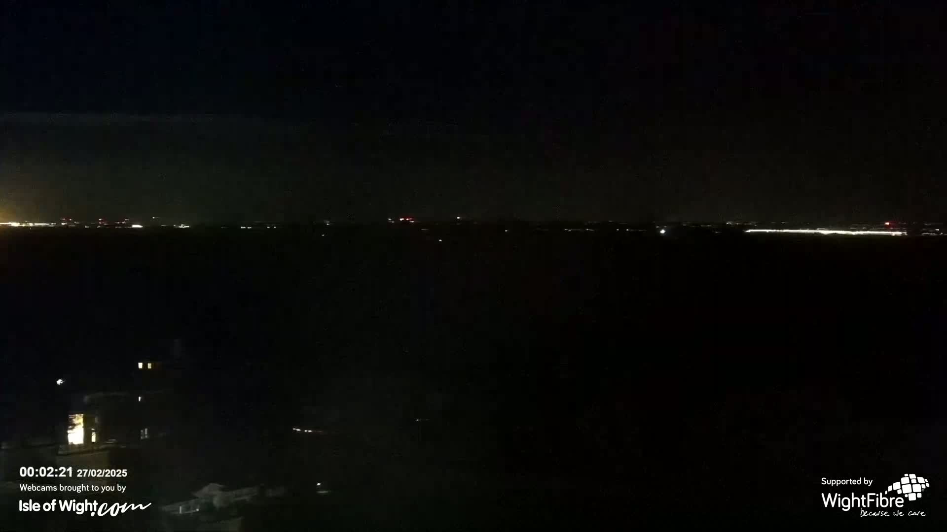 A dark, nighttime scene shows a distant line of lights across a dark body of water, with a few faint lights closer to the viewer in the bottom left corner.