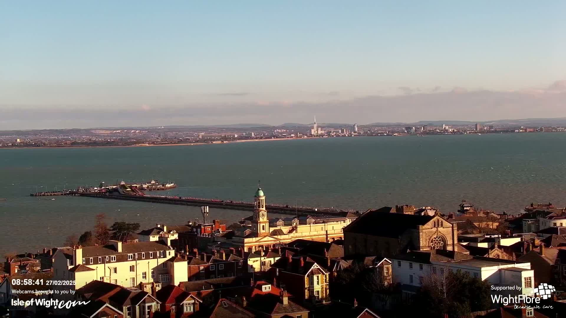 An aerial view of a coastal town on a sunny day, showing a pier extending into a calm body of water with a distant city skyline visible across the bay.
