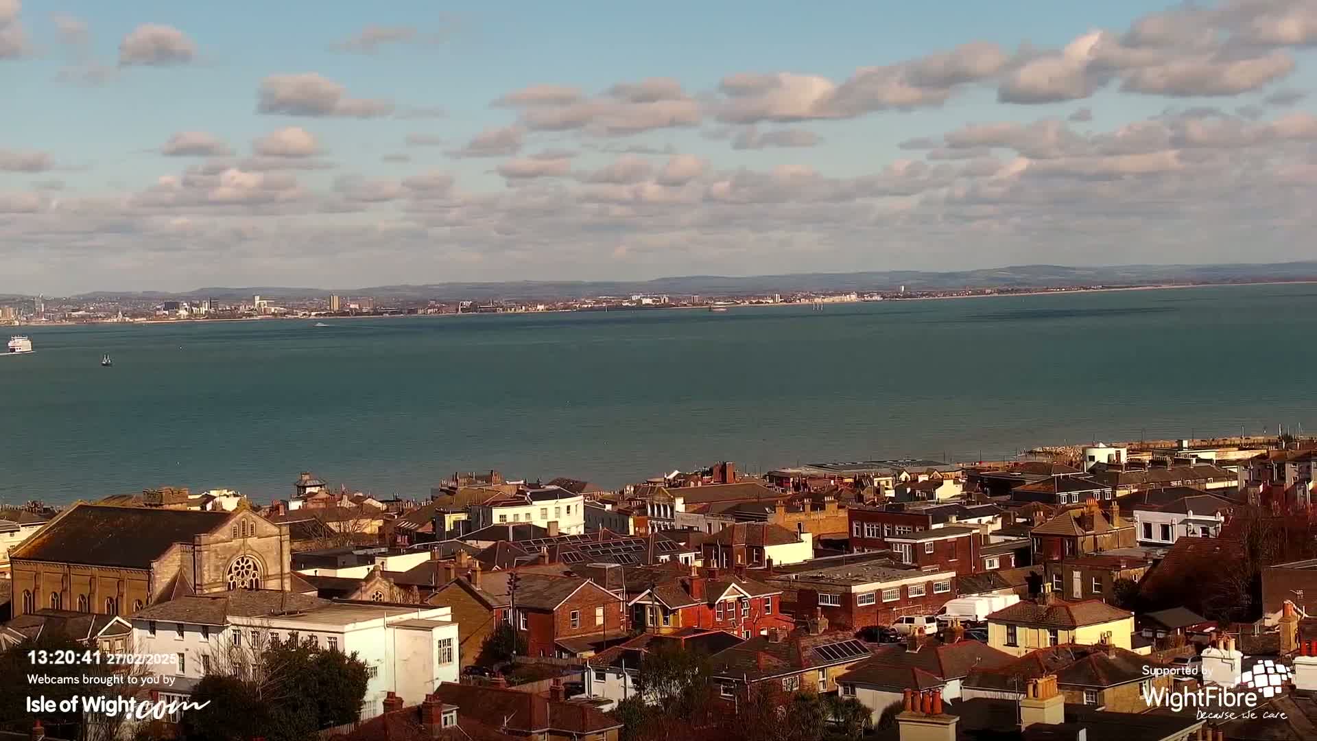 A coastal town with many brick buildings is seen from an elevated viewpoint, overlooking a calm body of water under a partly cloudy sky.