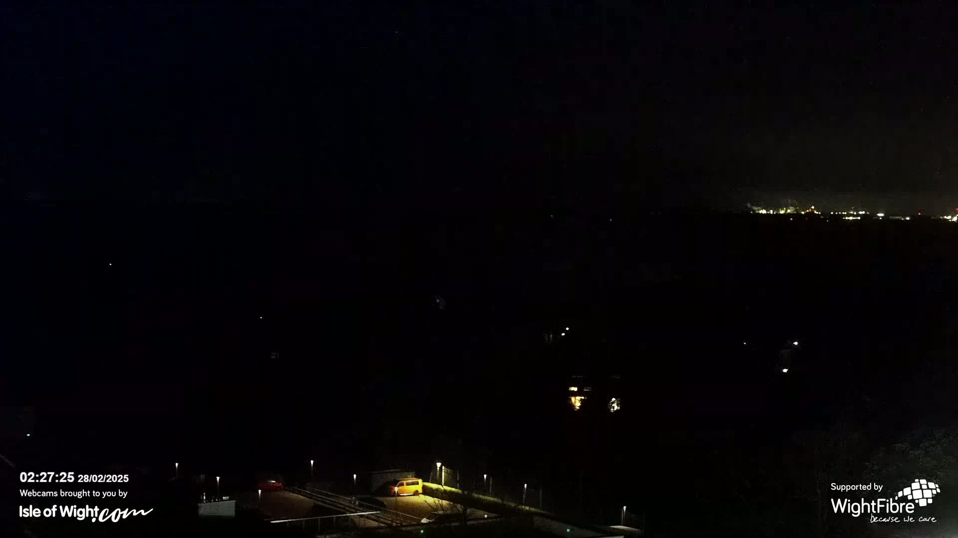 A nighttime, long shot of a dark landscape with a few distant lights and a small yellow vehicle on a road below.