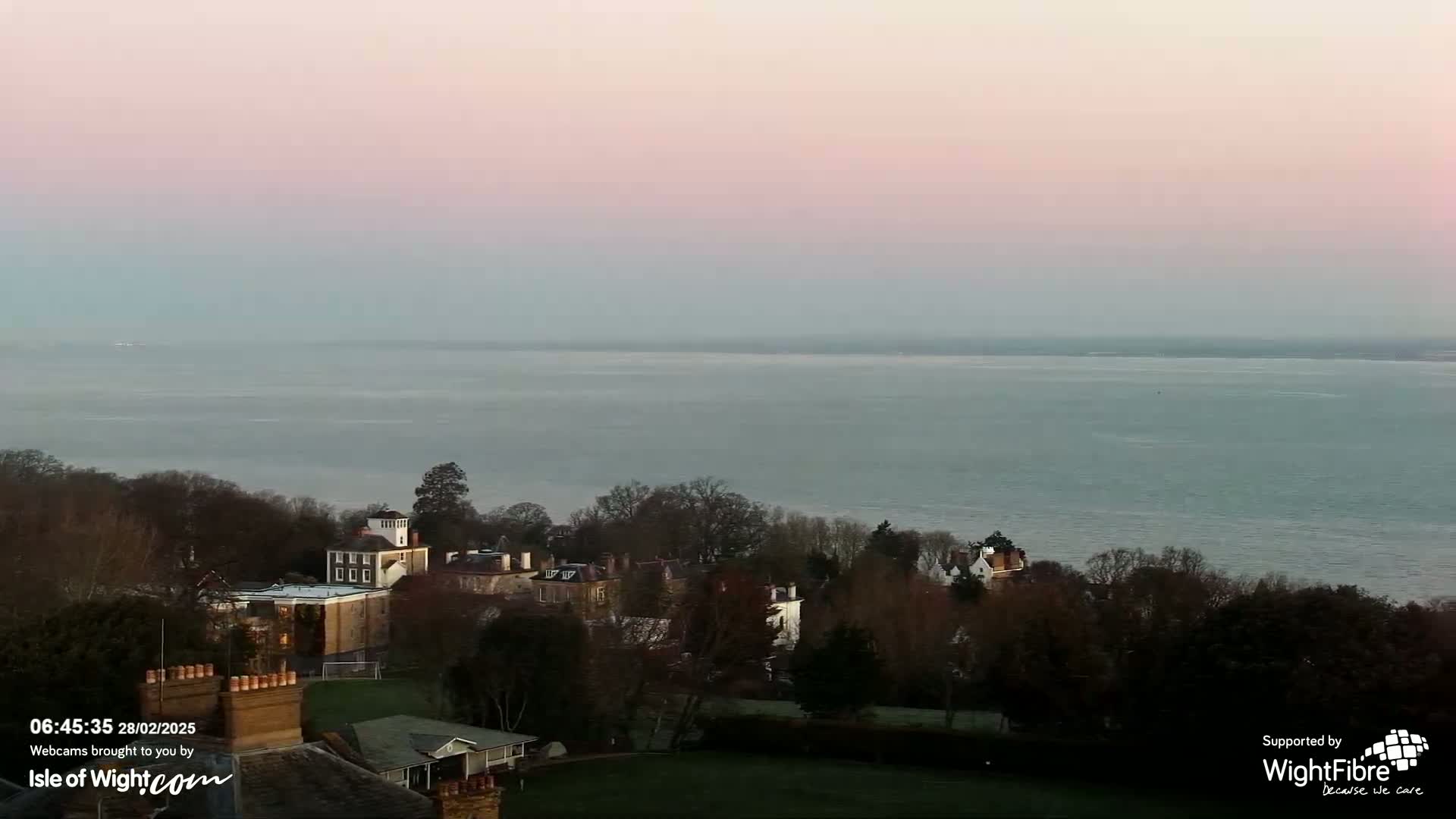 A calm, pale-pink sunrise illuminates a large body of water behind a cluster of buildings nestled among bare winter trees.