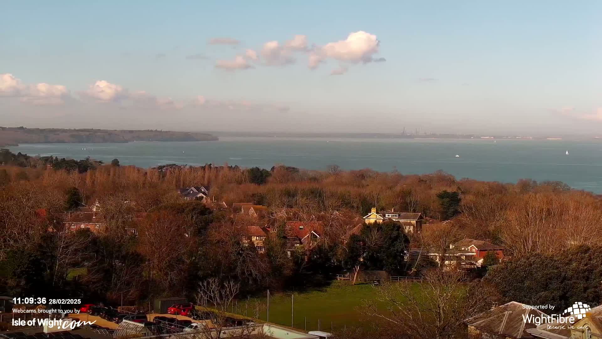 A panoramic view shows a residential area nestled among bare winter trees overlooking a calm body of water under a partly cloudy sky.