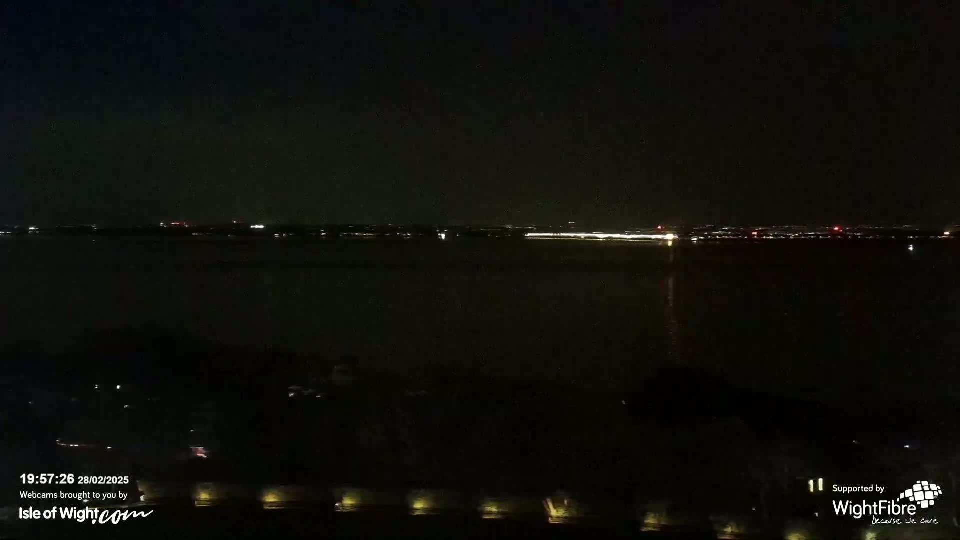A nighttime view of a dark body of water reflecting a distant line of city lights under a clear, dark sky.