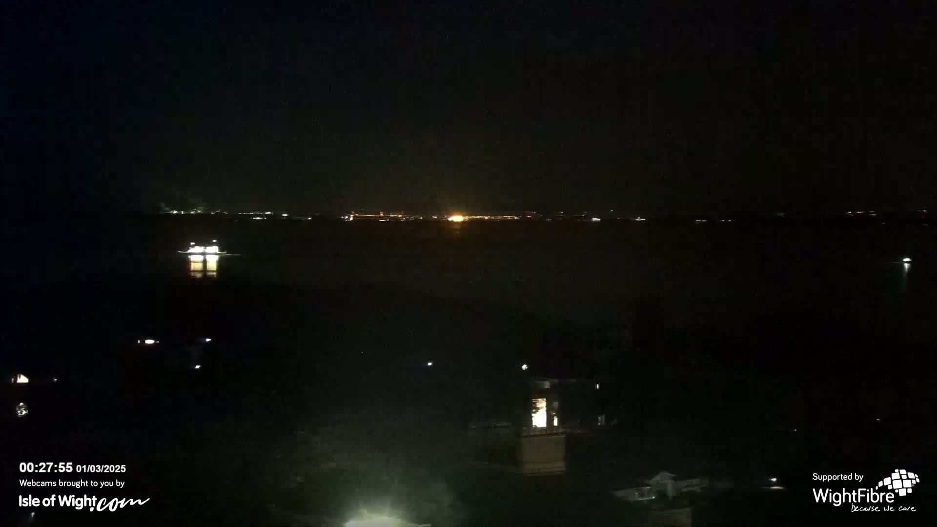 A nighttime view of a dark body of water with a few lights visible on the far shore and a single illuminated vessel in the foreground.