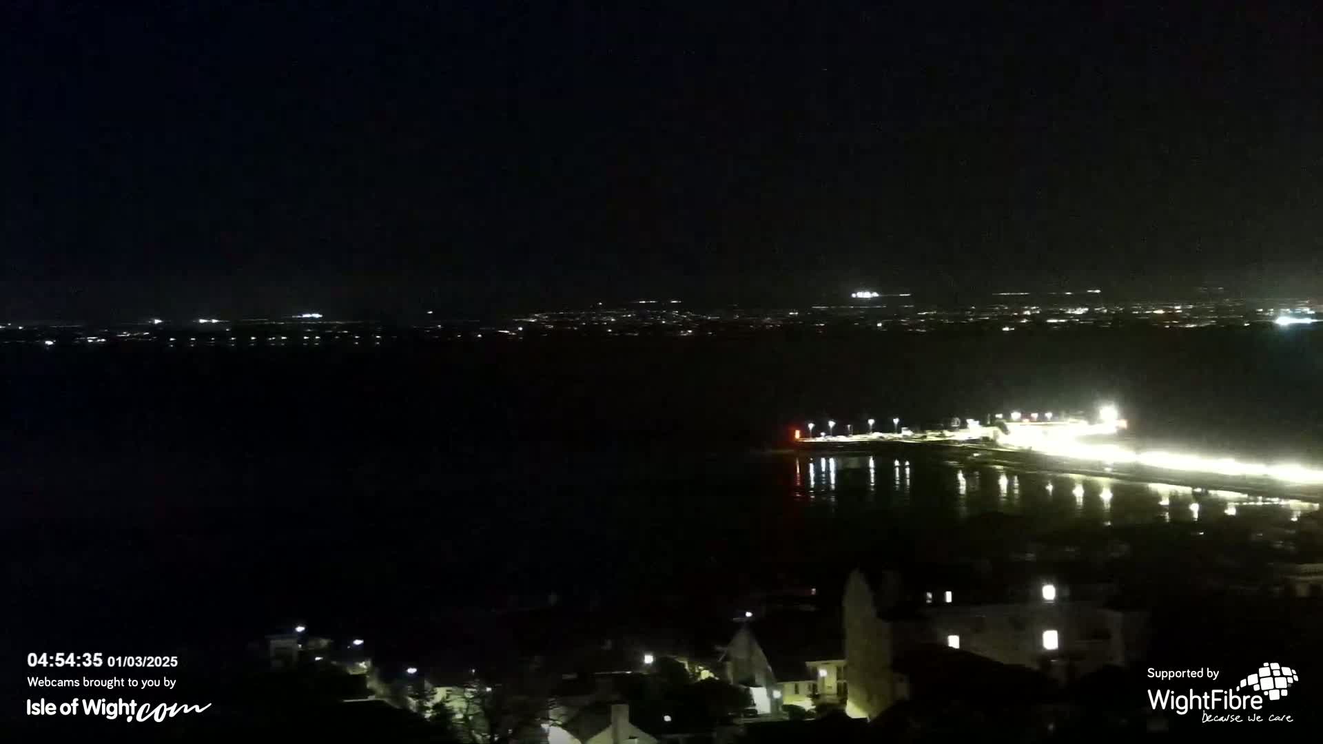 A nighttime view of a dark body of water reflecting lights from a distant shoreline and a town with illuminated buildings.