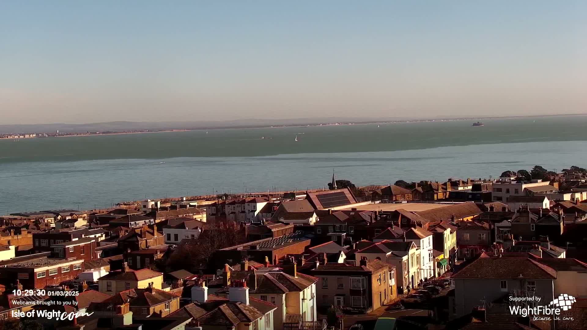 A coastal town overlooks a calm, wide body of water under a clear, sunny sky.