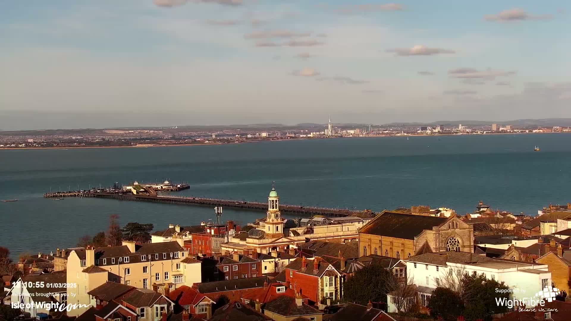A coastal town with red-tiled roofs and a clock tower overlooks a pier extending into a calm, blue body of water, with a distant city skyline visible under a mostly clear sky.