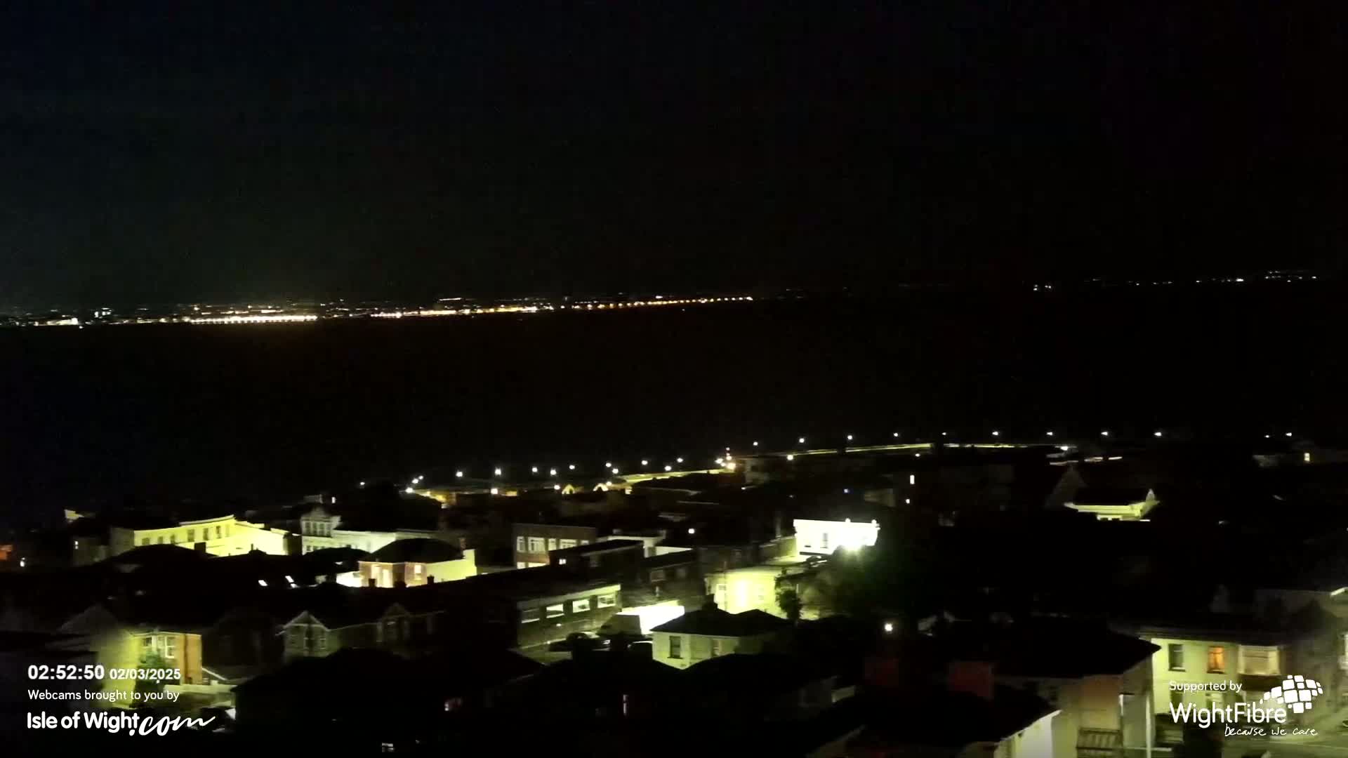 A nighttime view shows a town illuminated with streetlights, overlooking a dark body of water reflecting distant city lights under a clear sky.