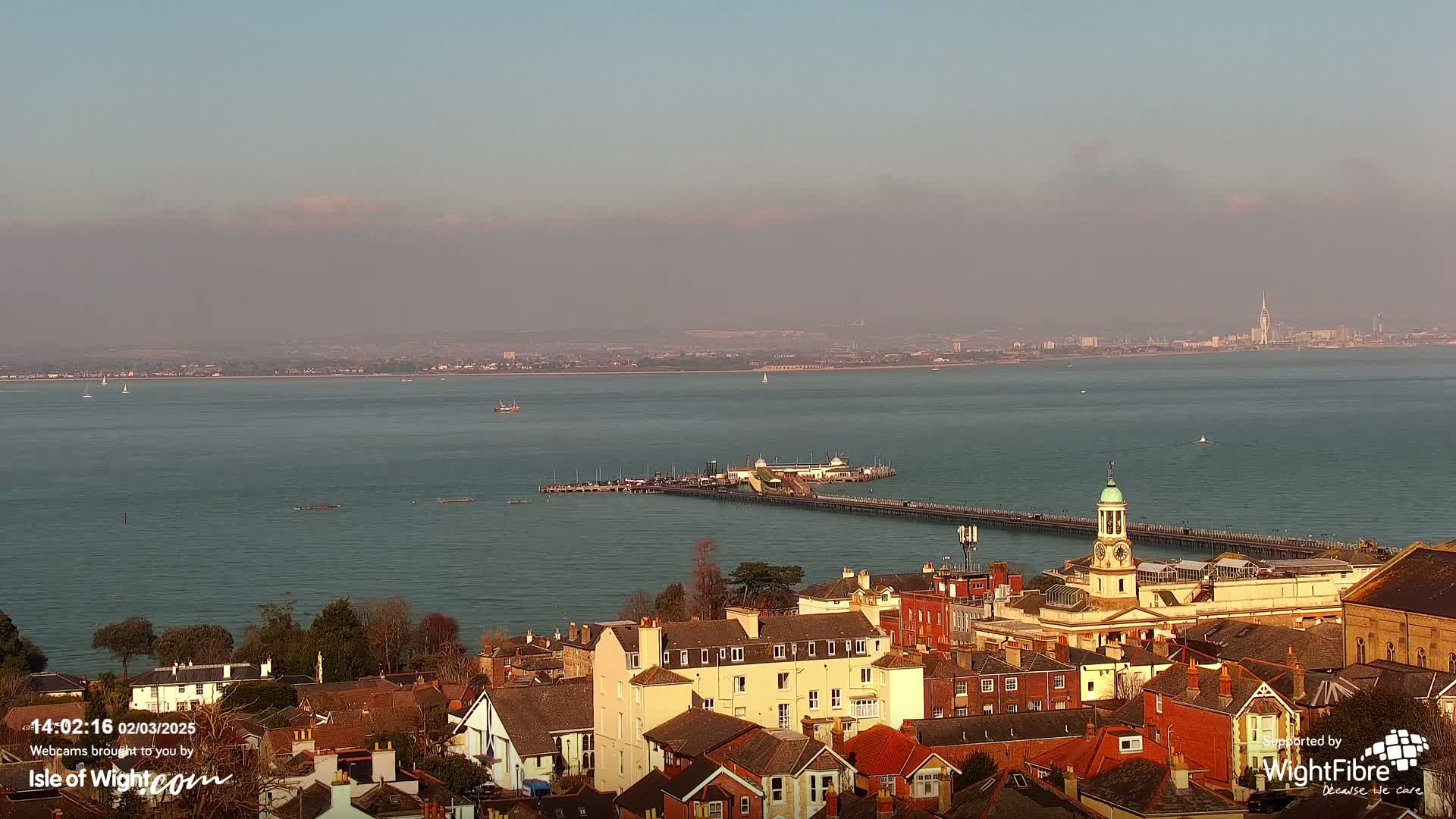 A hazy, sunny day overlooks a town with red-tiled roofs, a pier extending into a calm body of water, and a distant city skyline.