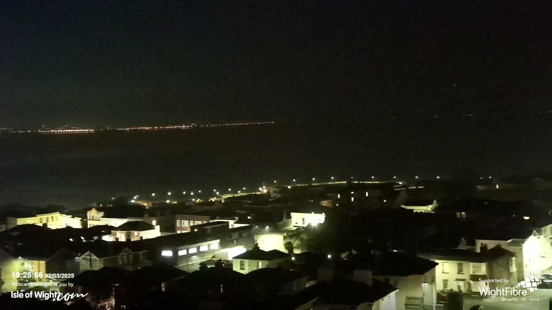 A nighttime view shows a coastal town illuminated by streetlights, overlooking a dark body of water with a distant line of lights on the opposite shore.