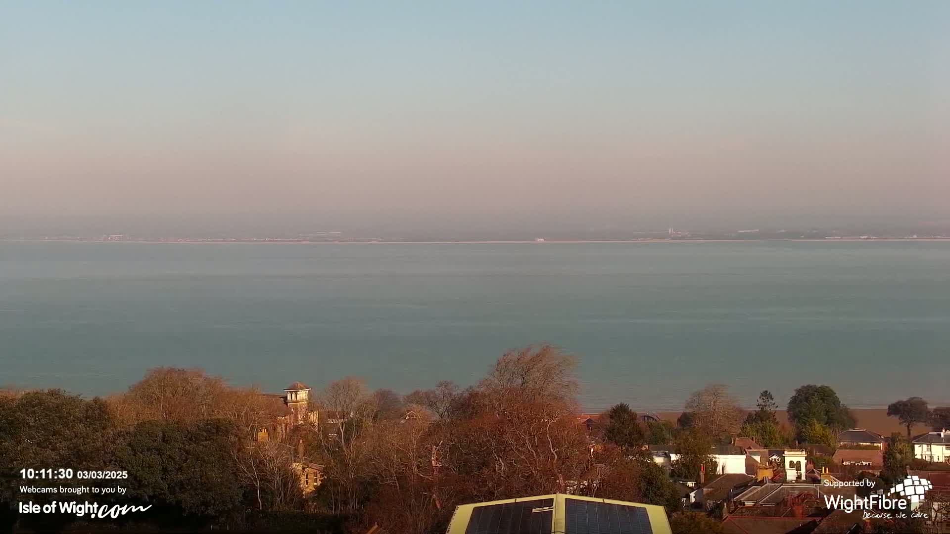 A calm, pale-blue body of water stretches to a hazy horizon under a clear, light-pink sky, overlooking a town of leafless trees and buildings.