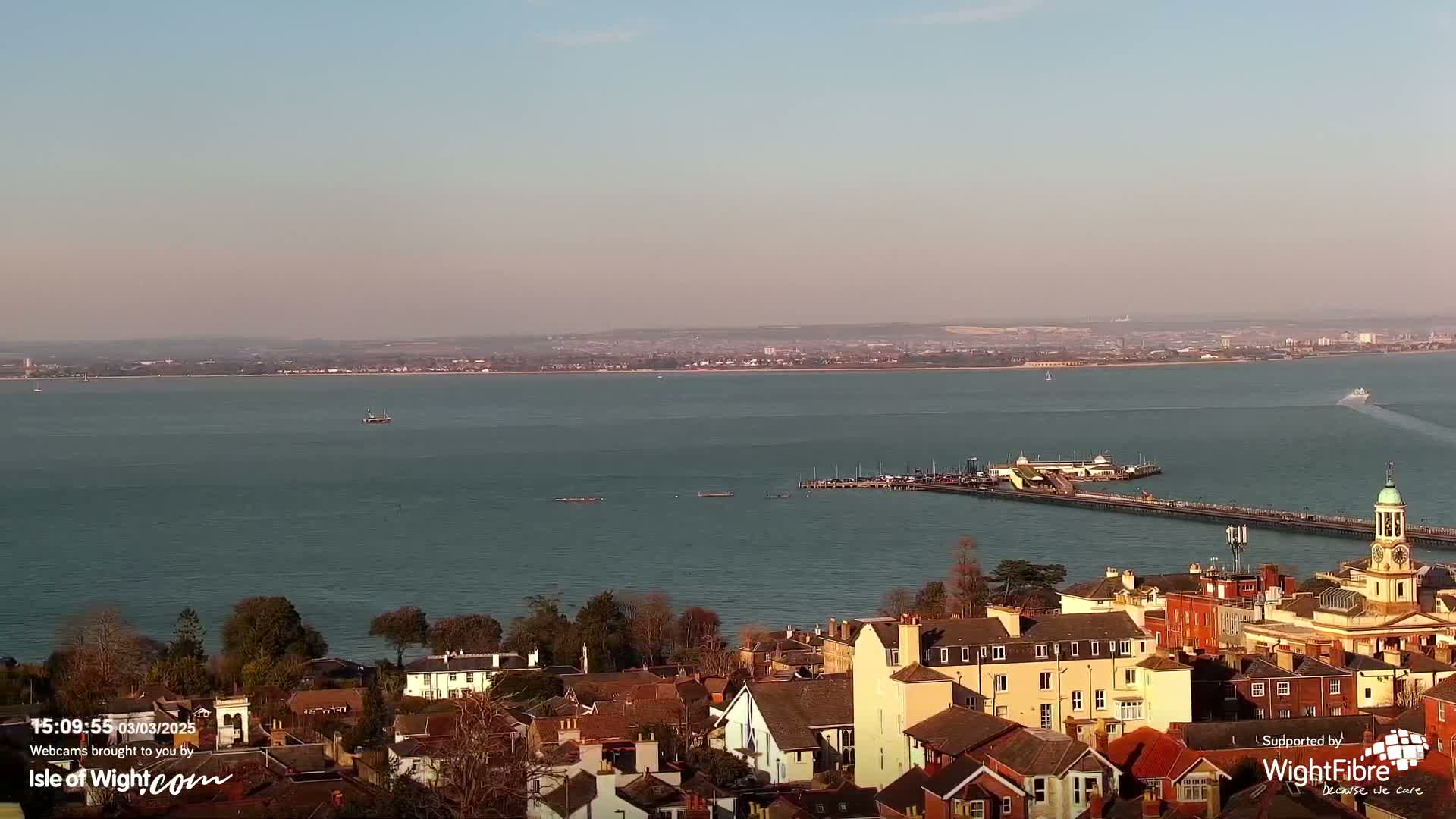 A coastal town with red-tiled roofs overlooks a calm body of water under a clear, pale sky, a long pier extending into the sea.