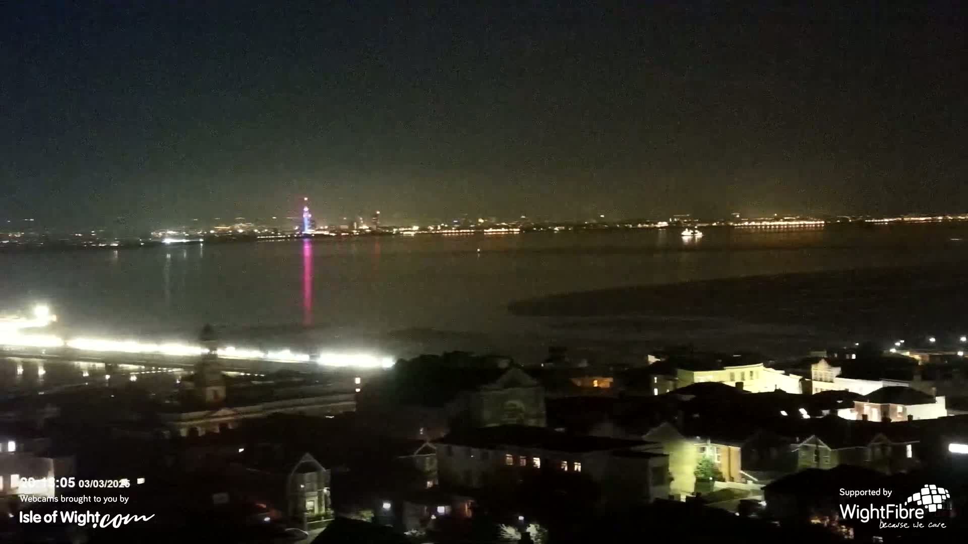 A nighttime view of a city across a body of water, with a distant, illuminated cityscape visible under a dark sky.