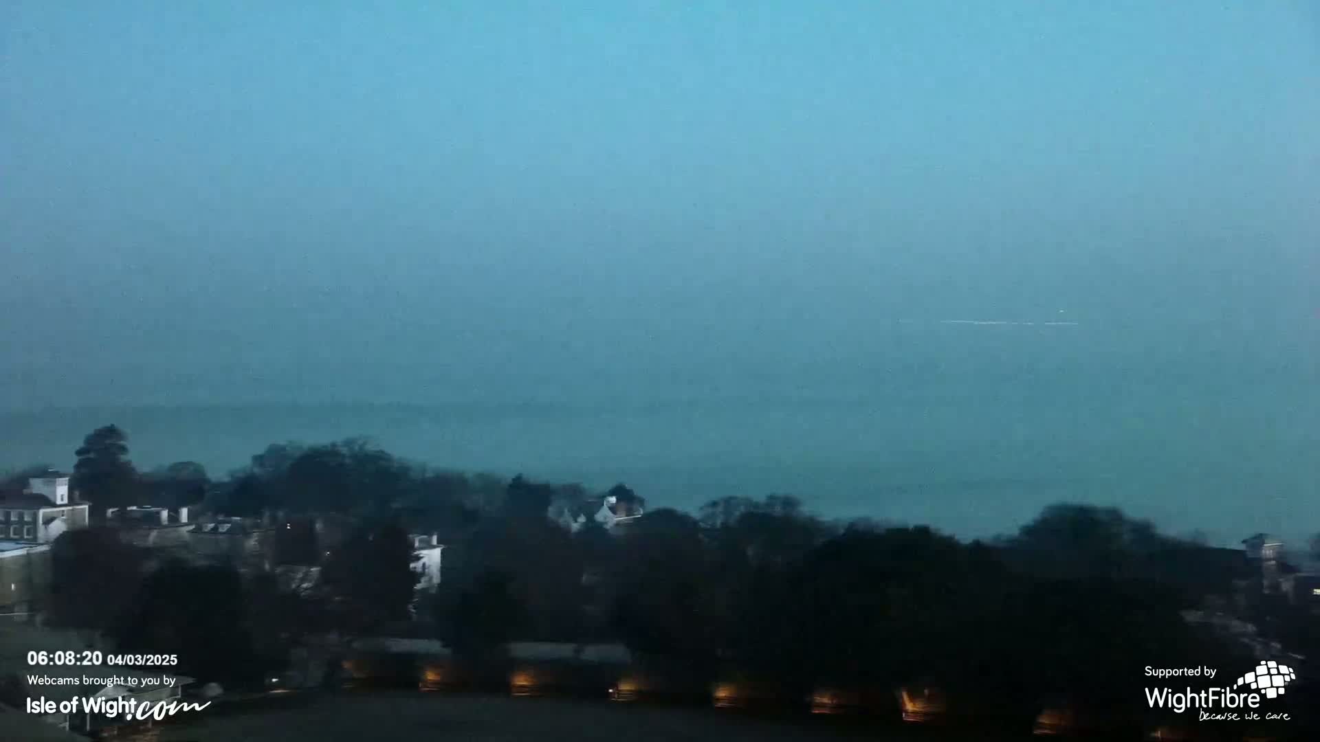 A hazy, bluish-gray morning reveals a distant body of water shrouded in mist, overlooking a line of dark trees and houses.