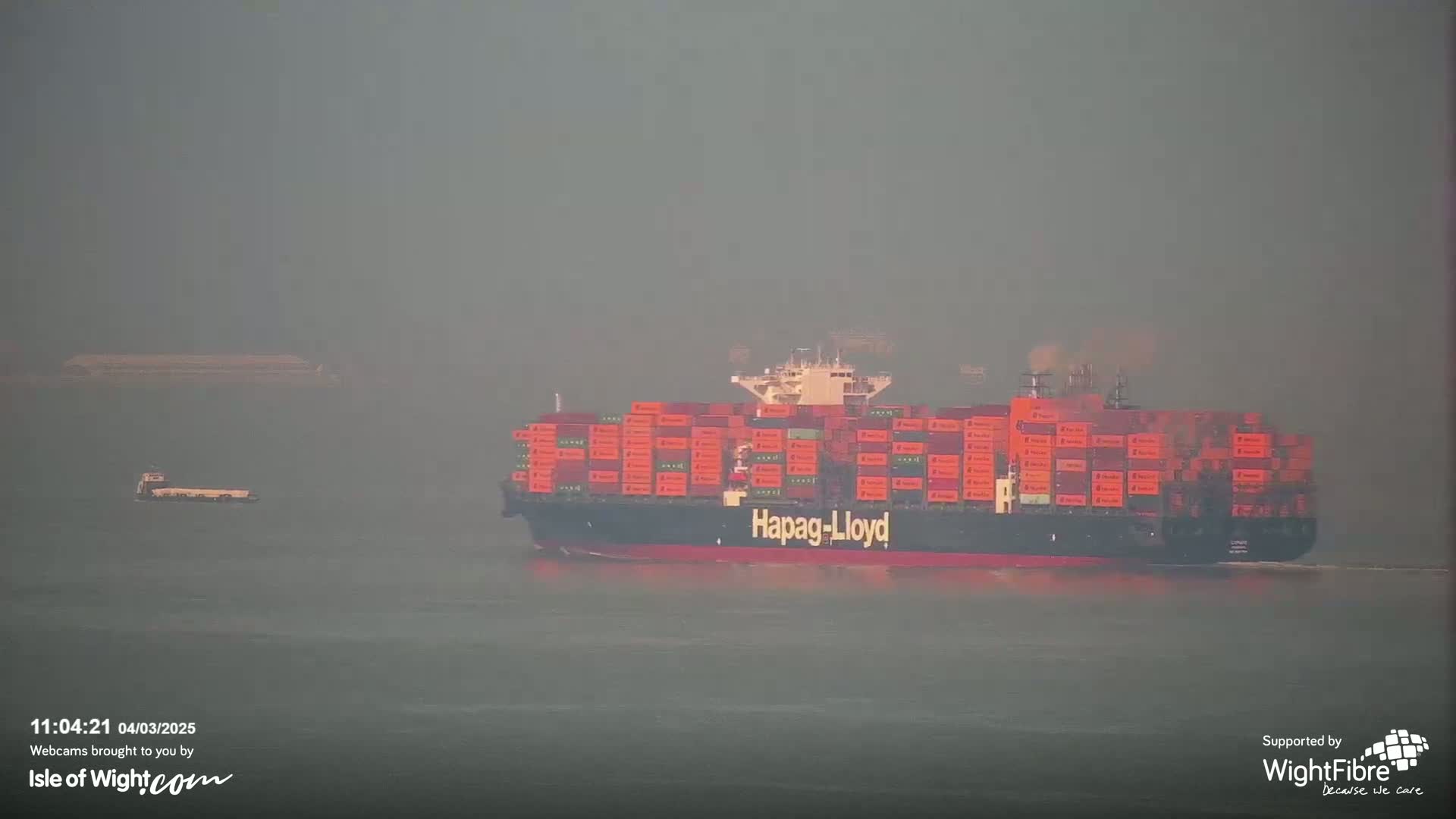 A large container ship, labeled Hapag-Lloyd, and a smaller vessel are visible on a hazy, overcast body of water.
