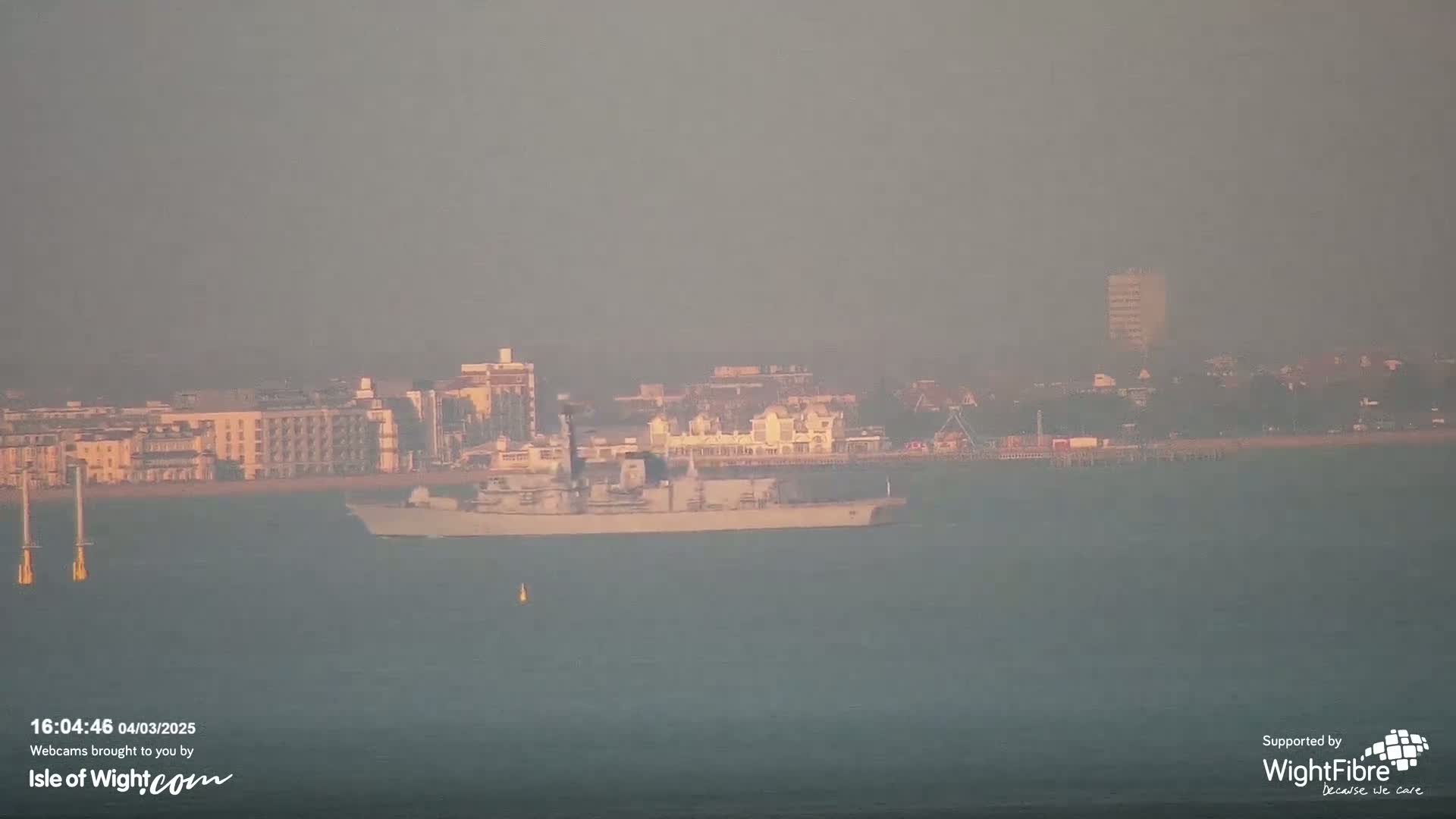 A hazy view shows a large warship on calm water off a distant coastline with buildings and a pier.
