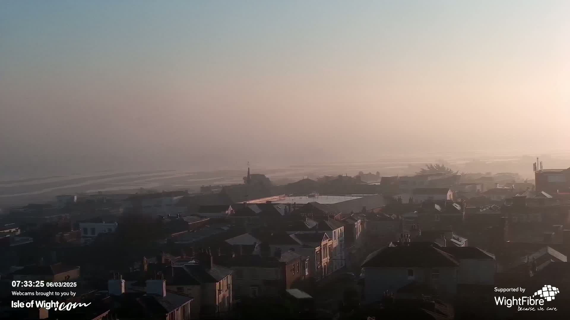 A hazy sunrise illuminates a town of tightly packed houses near a flat, misty coastline.