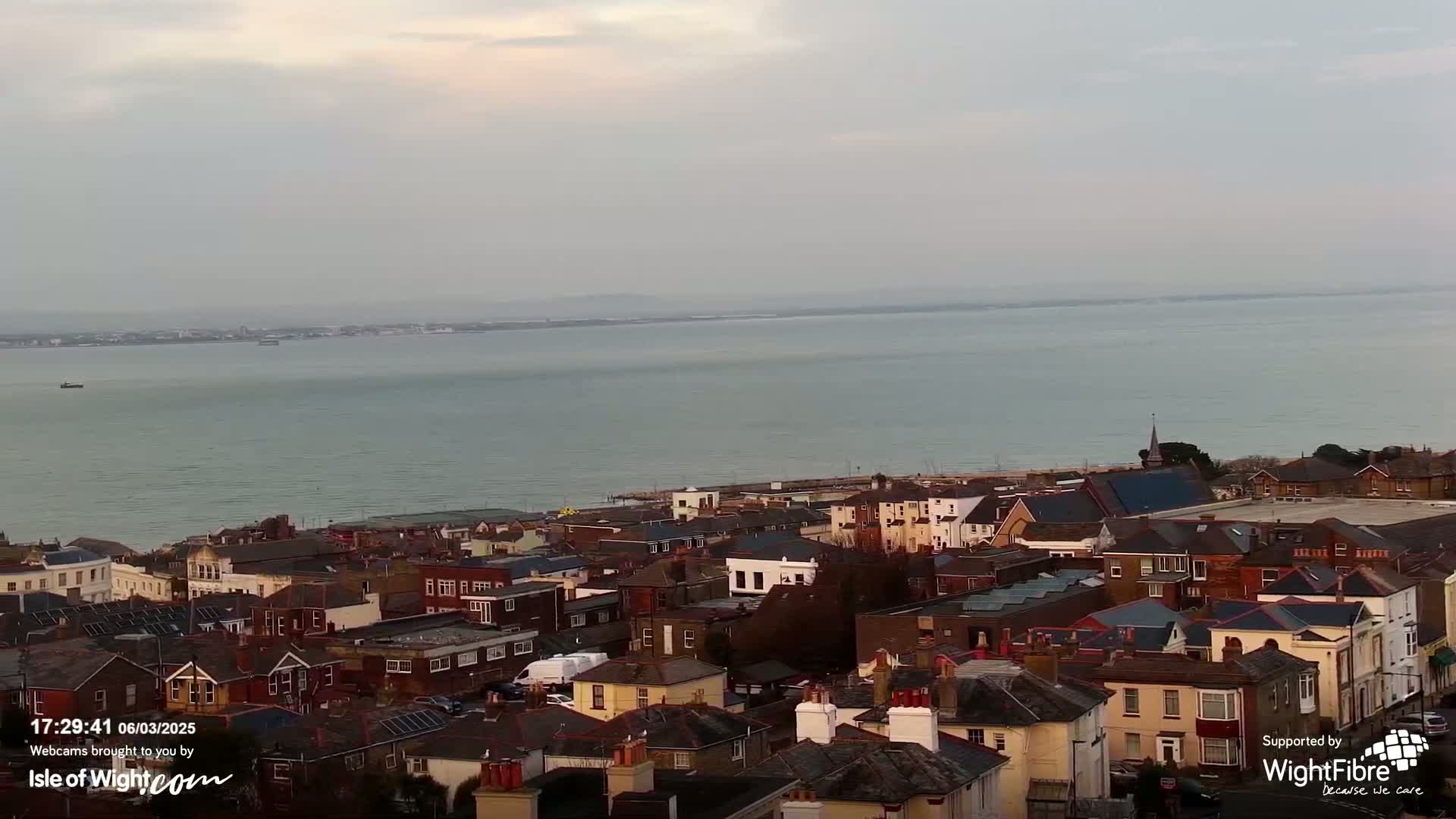 An aerial view shows a town of low-rise buildings next to a calm body of water under an overcast sky.